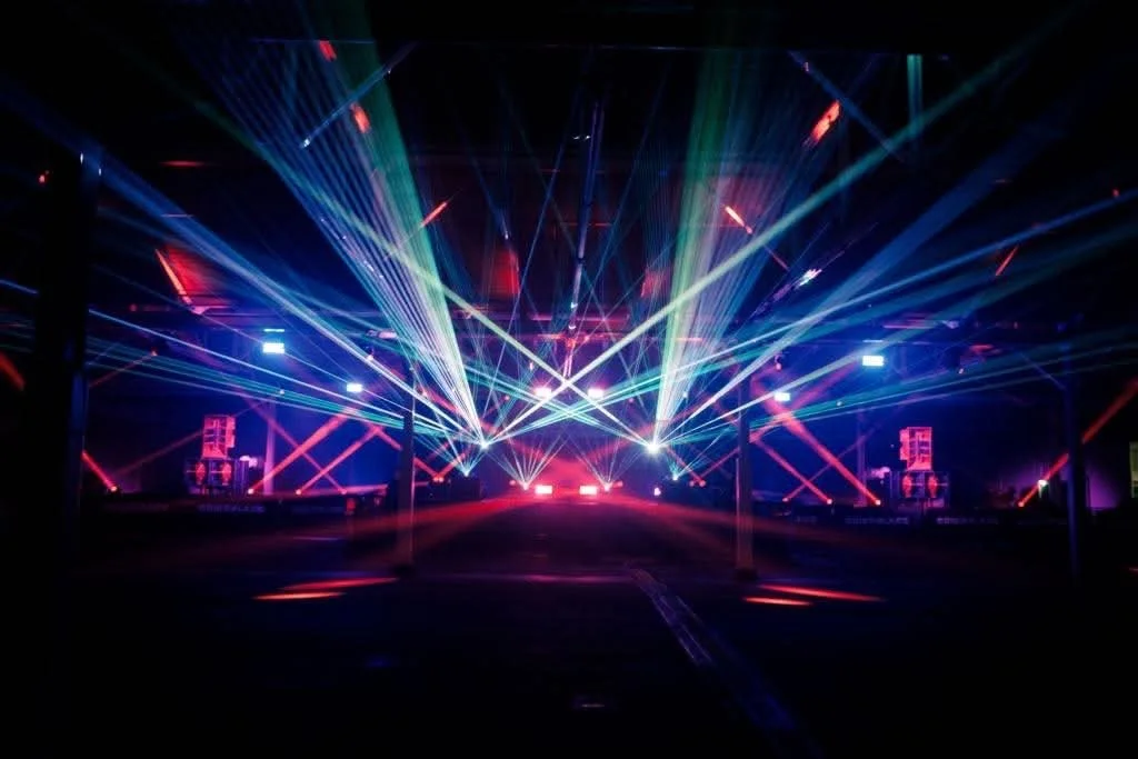 A dark indoor space with colorful laser lights and beams, likely at a concert or nightclub, creating a vibrant atmosphere.