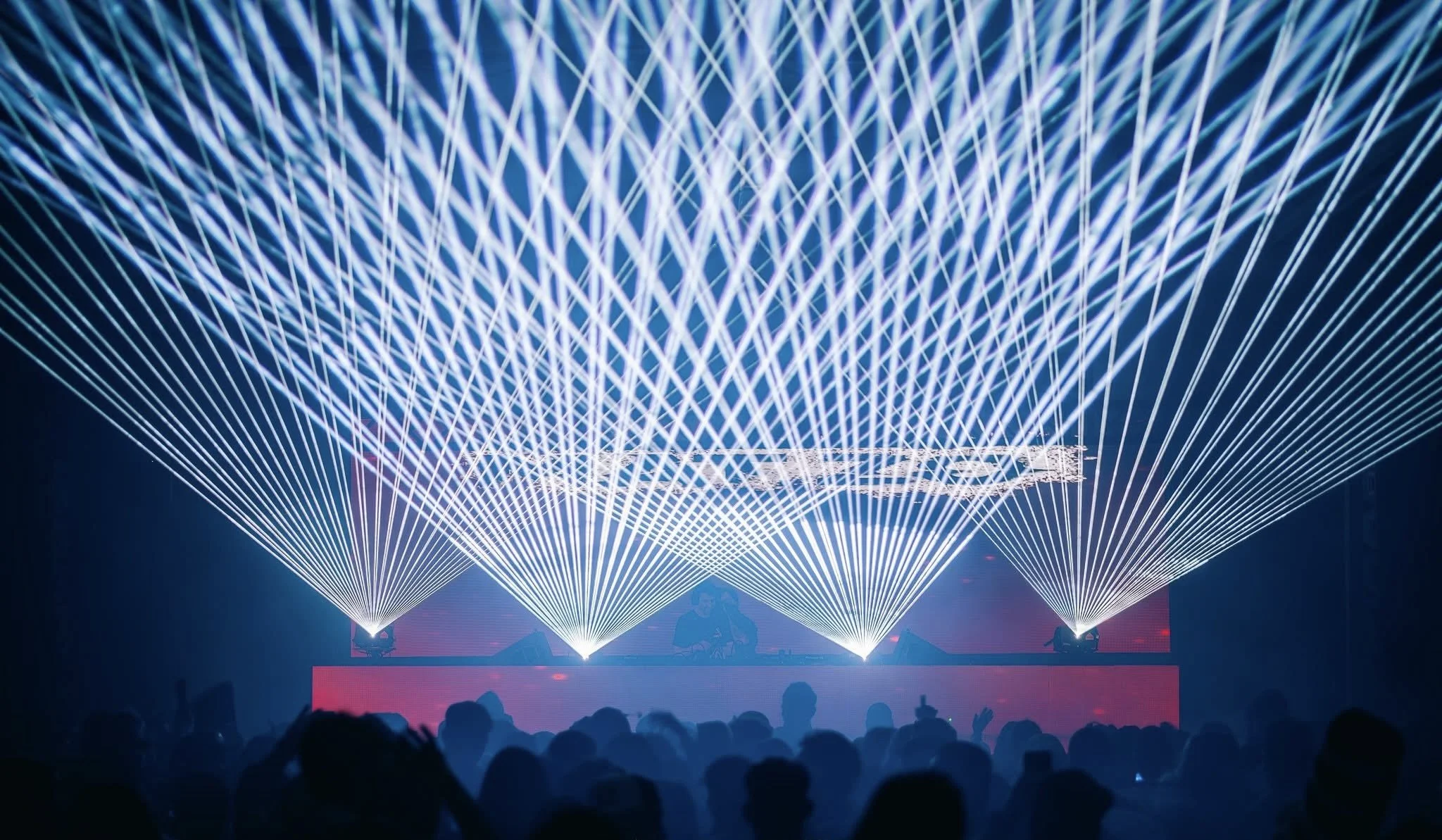 A light show with blue and white laser beams forming a geometric pattern above a crowd at a concert or event.