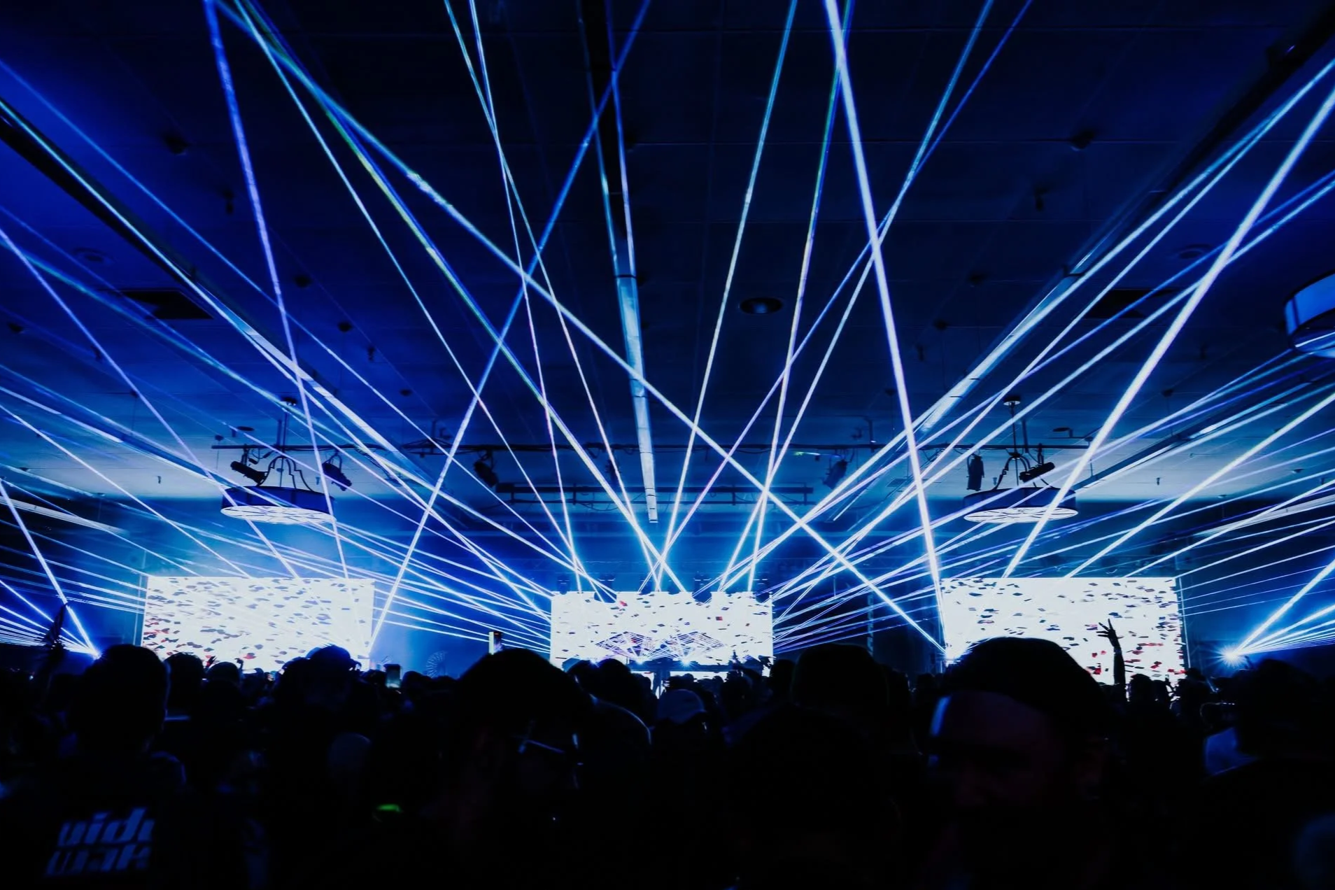 Dark room filled with an audience watching a concert or event with blue laser lights and LED screens displaying abstract visuals.