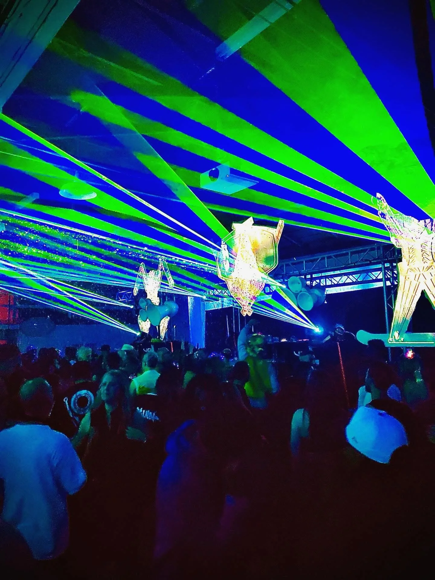 Crowd attending a concert or nightclub with green and blue laser lights, illuminated balloons, and decorative objects hanging from the ceiling.