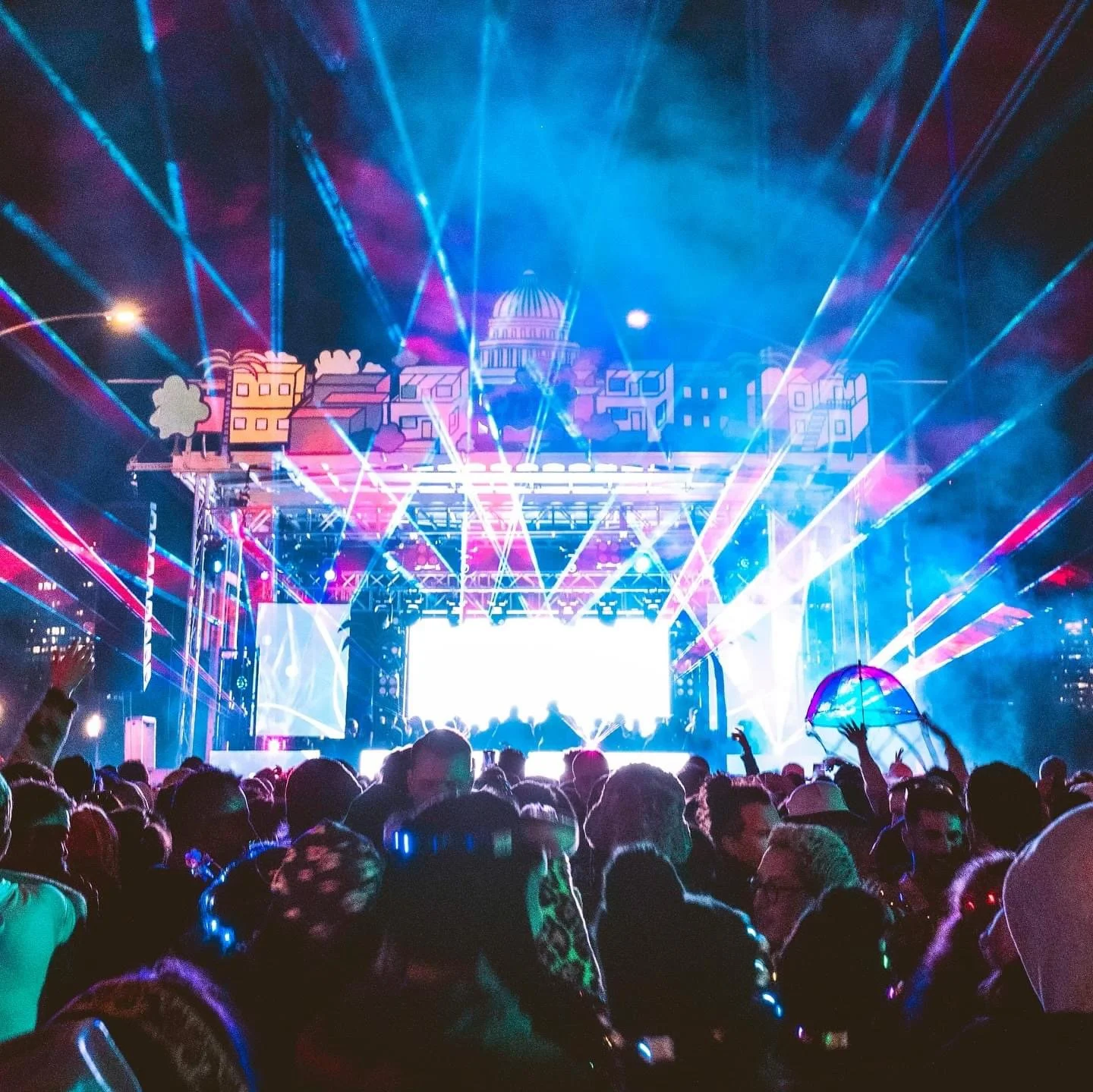 A large crowd attending a concert or festival at night, with colorful laser lights, a stage with LED screens, and artwork of city buildings and a dome-shaped structure in the background.