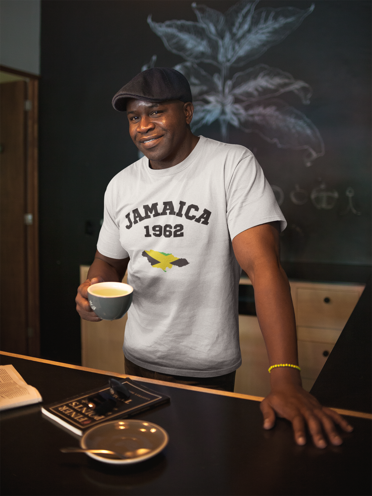 Man in a cafe wearing a "Jamaica 1962" t-shirt, holding a coffee cup and smiling, with a hat on his head.