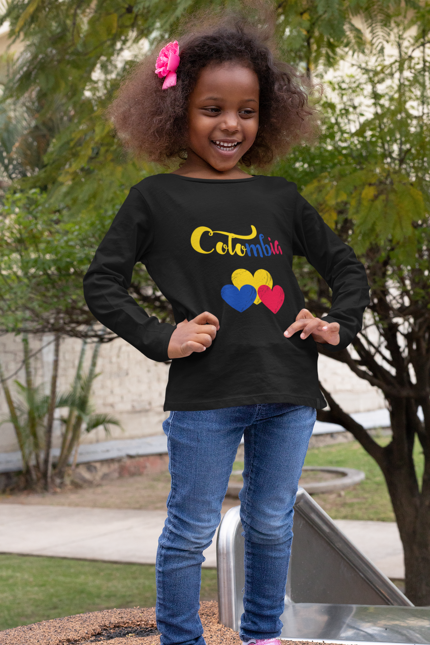 Young girl with curly hair, smiling, wearing a black long-sleeve shirt with the word 'Colombia' and three colored hearts on it, standing outdoors next to a playground slide.