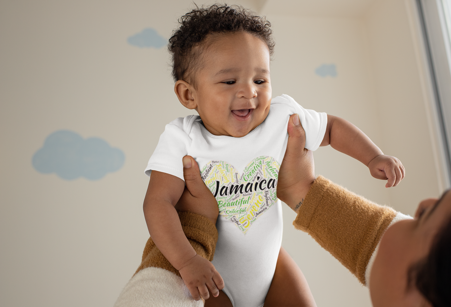 Smiling baby held up wearing a onesie with "Jamaica" and a colorful heart design, background with blue clouds.