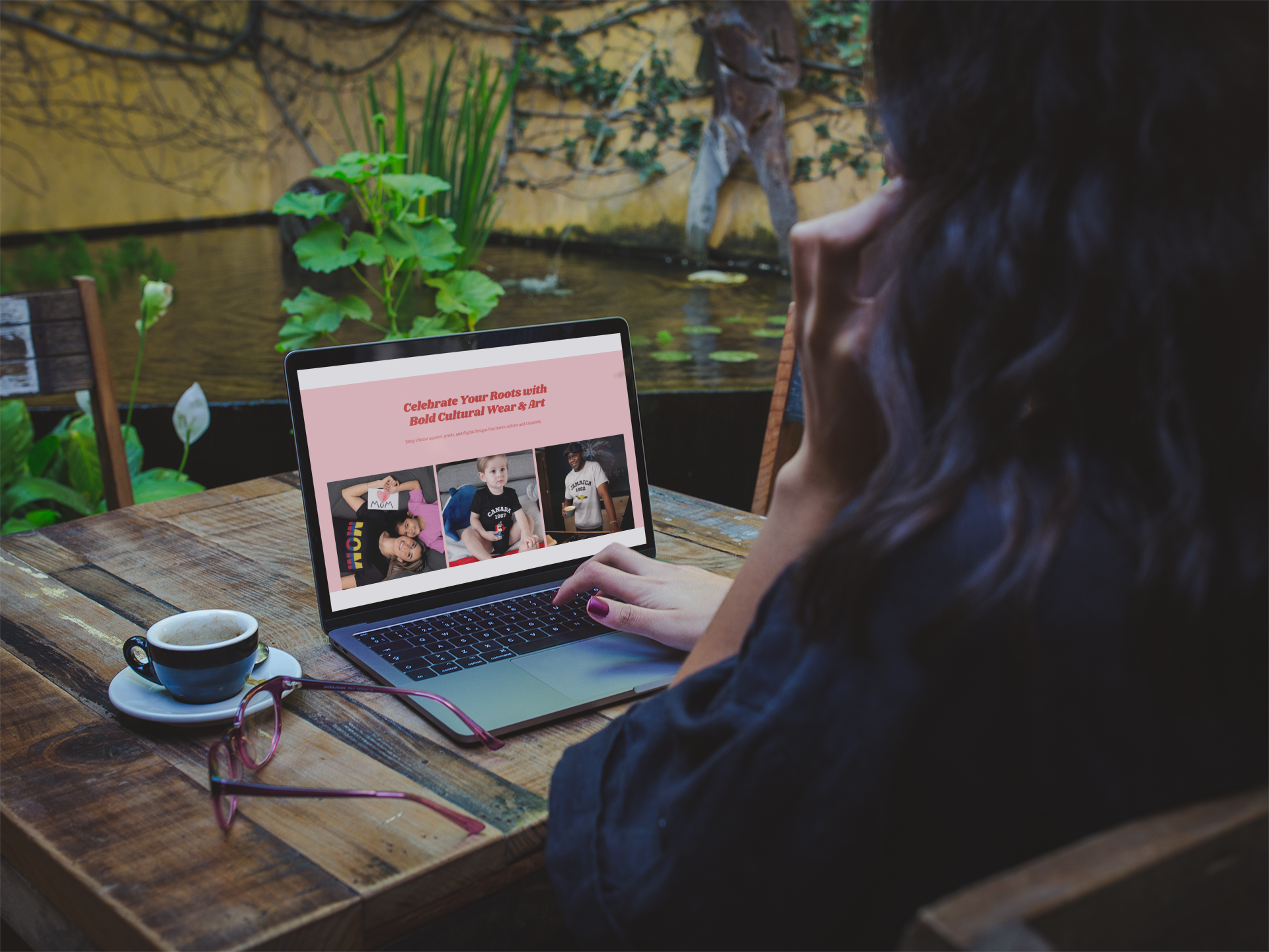 A woman sitting at a wooden table, looking at a laptop with a website for celebrating cultural wear and art, with a cup of coffee and pink glasses on the table, near a pond with plants and a yellow wall in the background.