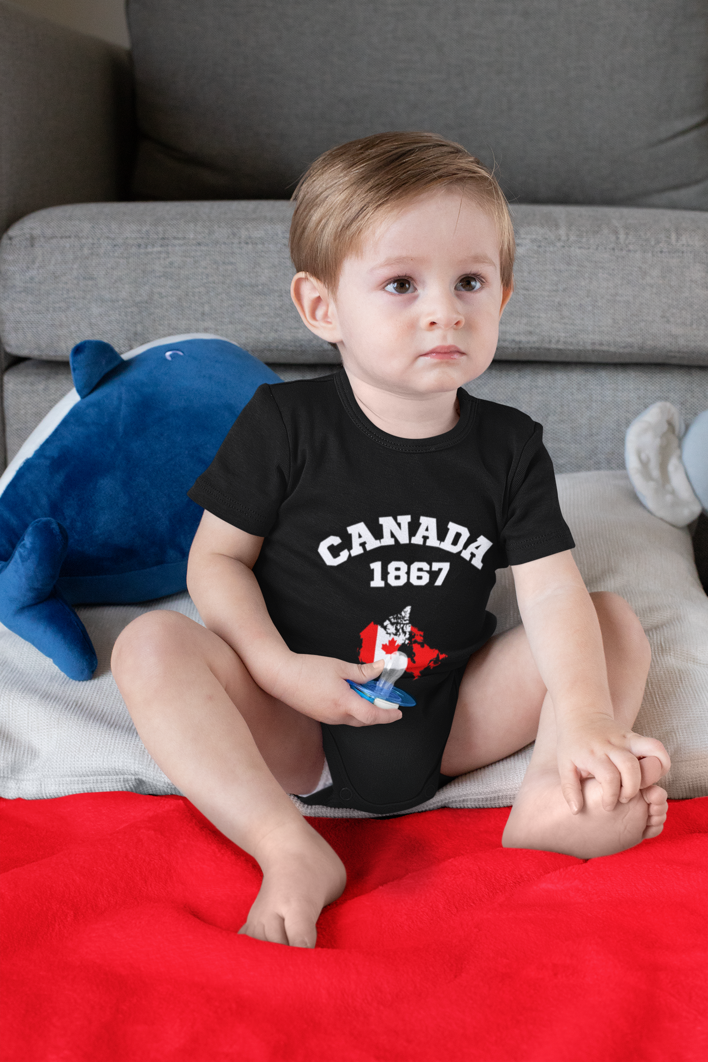 A young boy sitting on a bed with a red blanket, wearing a black shirt that says 'Canada 1867' with a maple leaf graphic. There is a plush blue whale toy behind him and a light-colored pillow to his right.
