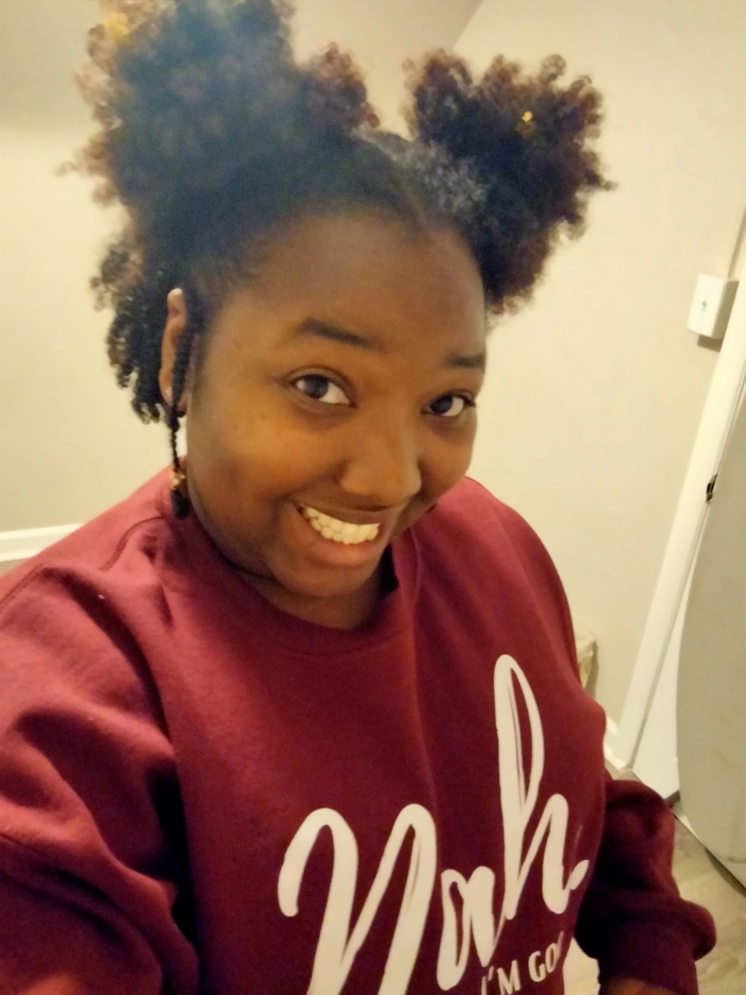 A woman smiling with natural hair styled in two large puffs, wearing a maroon sweatshirt that says 'Wow I'm Good' on the front.