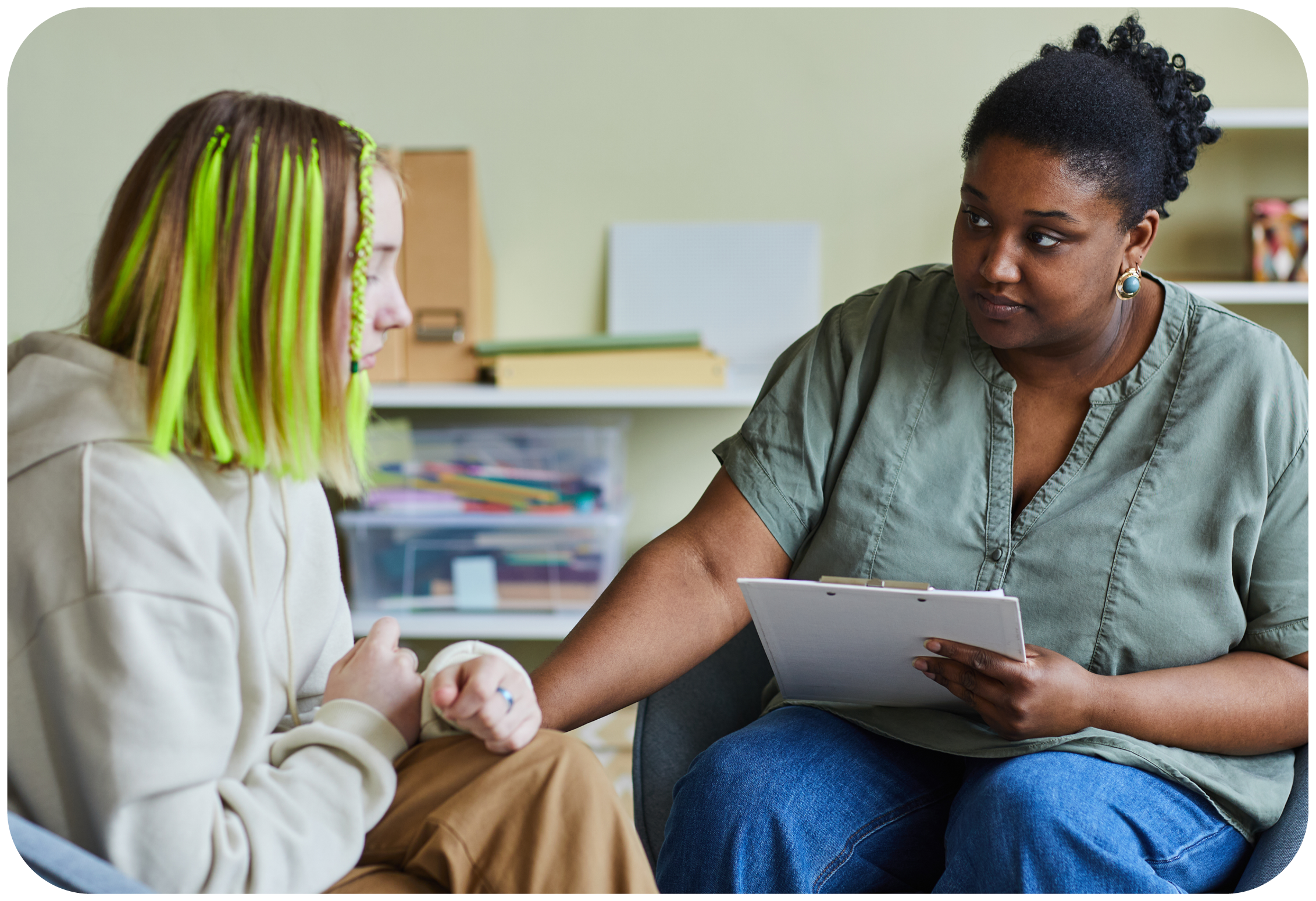 A woman with natural curly hair and earrings talking to a young girl with long neon green and brown braids and wearing a cream hoodie, while the woman holds a clipboard.