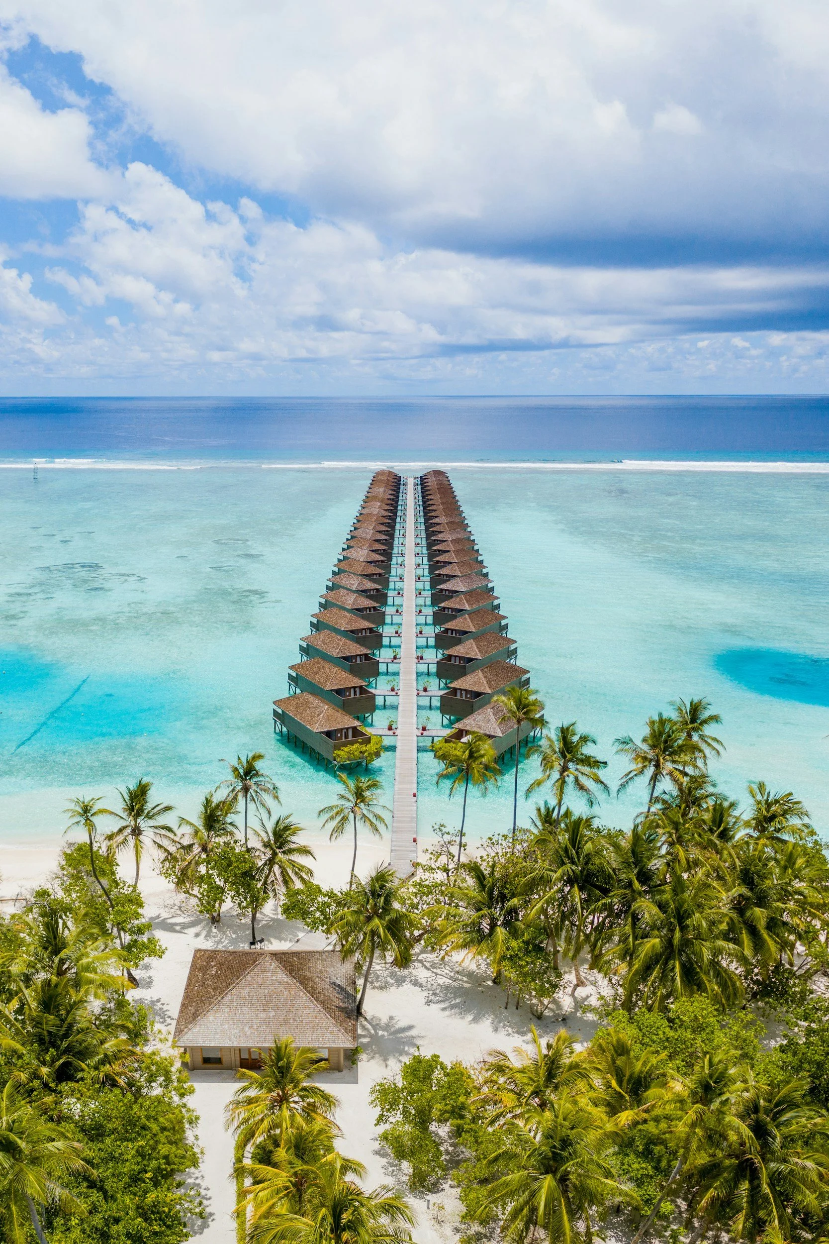 Overwater bungalows extending into turquoise ocean, connected by a walkway, with a sandy beach and palm trees in the foreground, and a cloudy sky in the background.