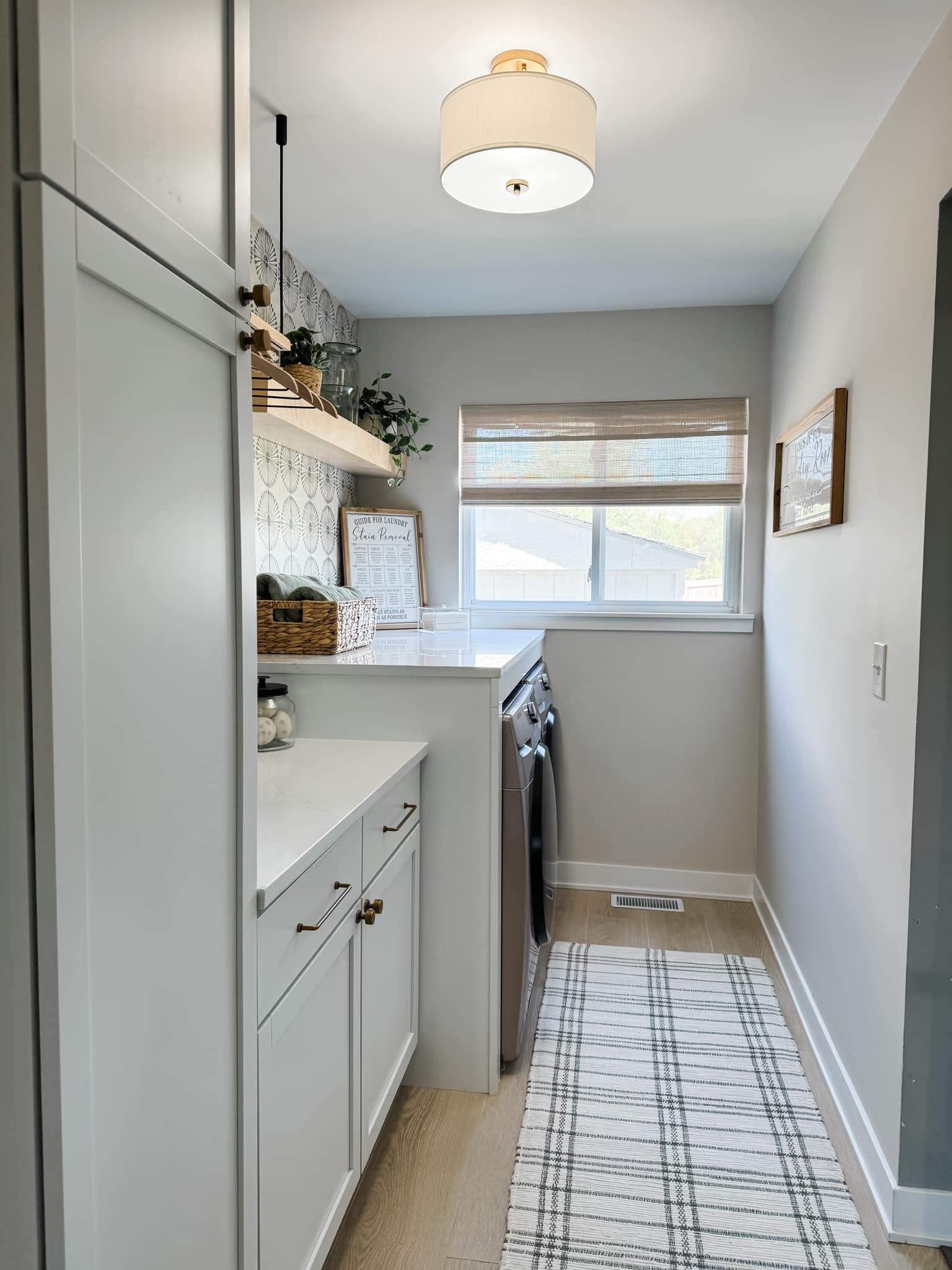 Stylish laundry room with white cabinets, washer and dryer, patterned wallpaper, floating shelf with decorative items, and a woven rug on light wood floor.