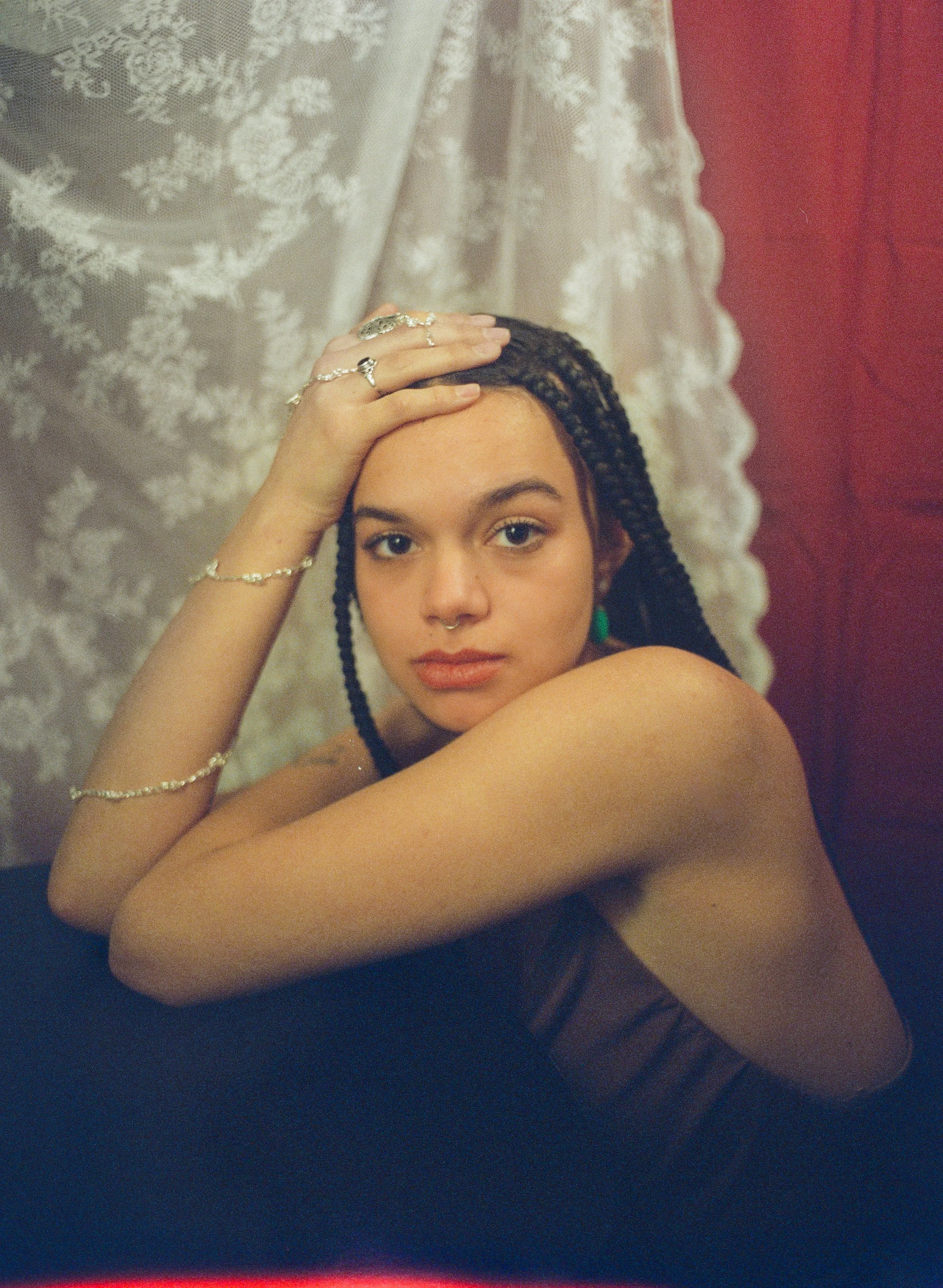 A woman with braided hair, wearing jewelry, poses with her arm resting on her head against a lace and red fabric backdrop.