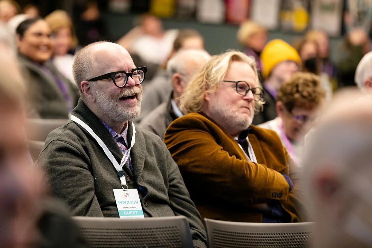 Crowd of people attending a conference, with two men in the foreground, one laughing and the other looking serious, both wearing glasses and conference badges.