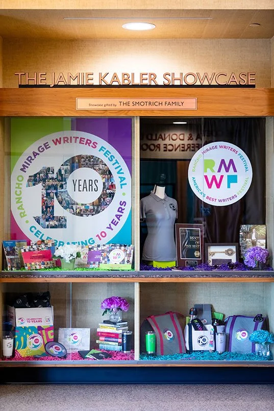 Display case celebrating the 10th anniversary of the Mirage Writers Festival, featuring a large poster with the festival logo, books, flowers, and memorabilia.