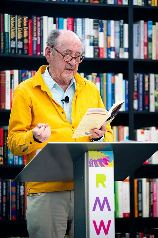 An elderly man with glasses and gray hair, wearing a yellow jacket, standing at a podium in front of a bookshelf filled with colorful books, reading from a book.