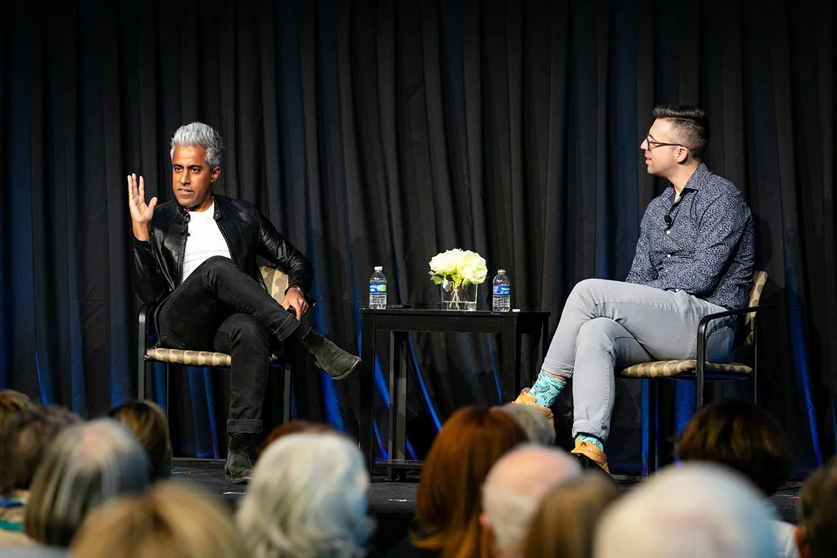 Two men seated on a stage engaged in conversation, with an audience in front and a black curtain backdrop. One man has gray hair and is wearing a black leather jacket, white shirt, and black pants, gesturing with his hand. The other man has dark hair