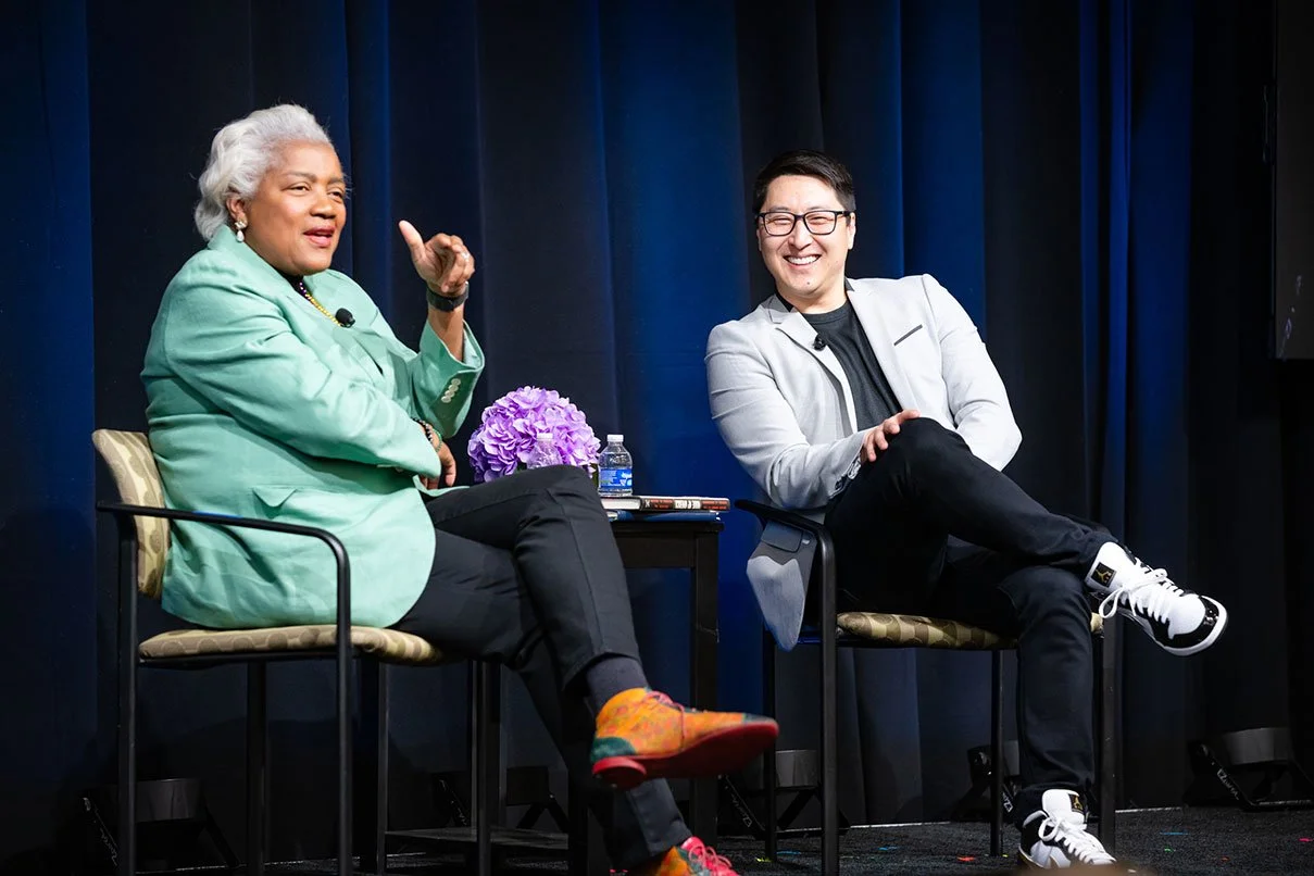 An elderly woman with gray hair and a young man with glasses seated on stage, smiling and talking. The woman is wearing a green blazer and multicolored shoes, while the man wears a white jacket and black pants. A small table with purple flowers and a
