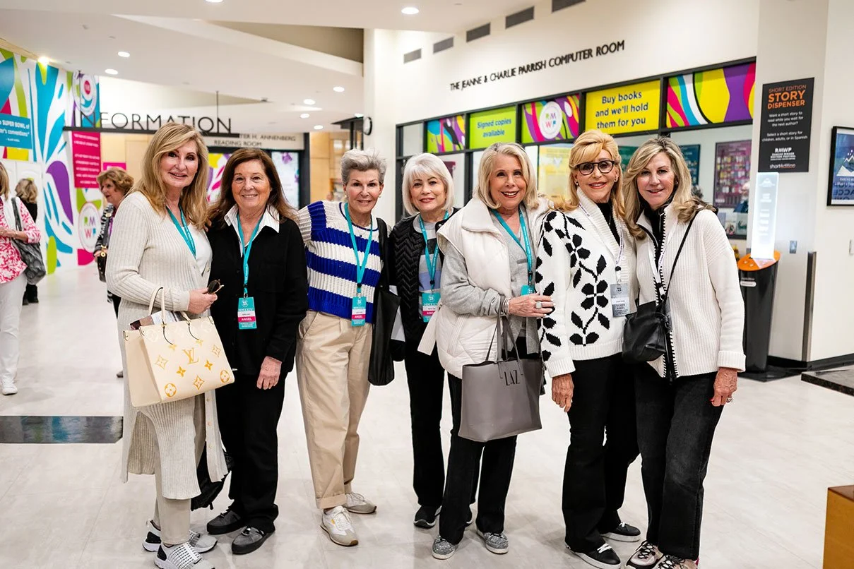 Group of seven older women standing together inside a modern building, smiling at the camera, with colorful signage and a digital display in the background.