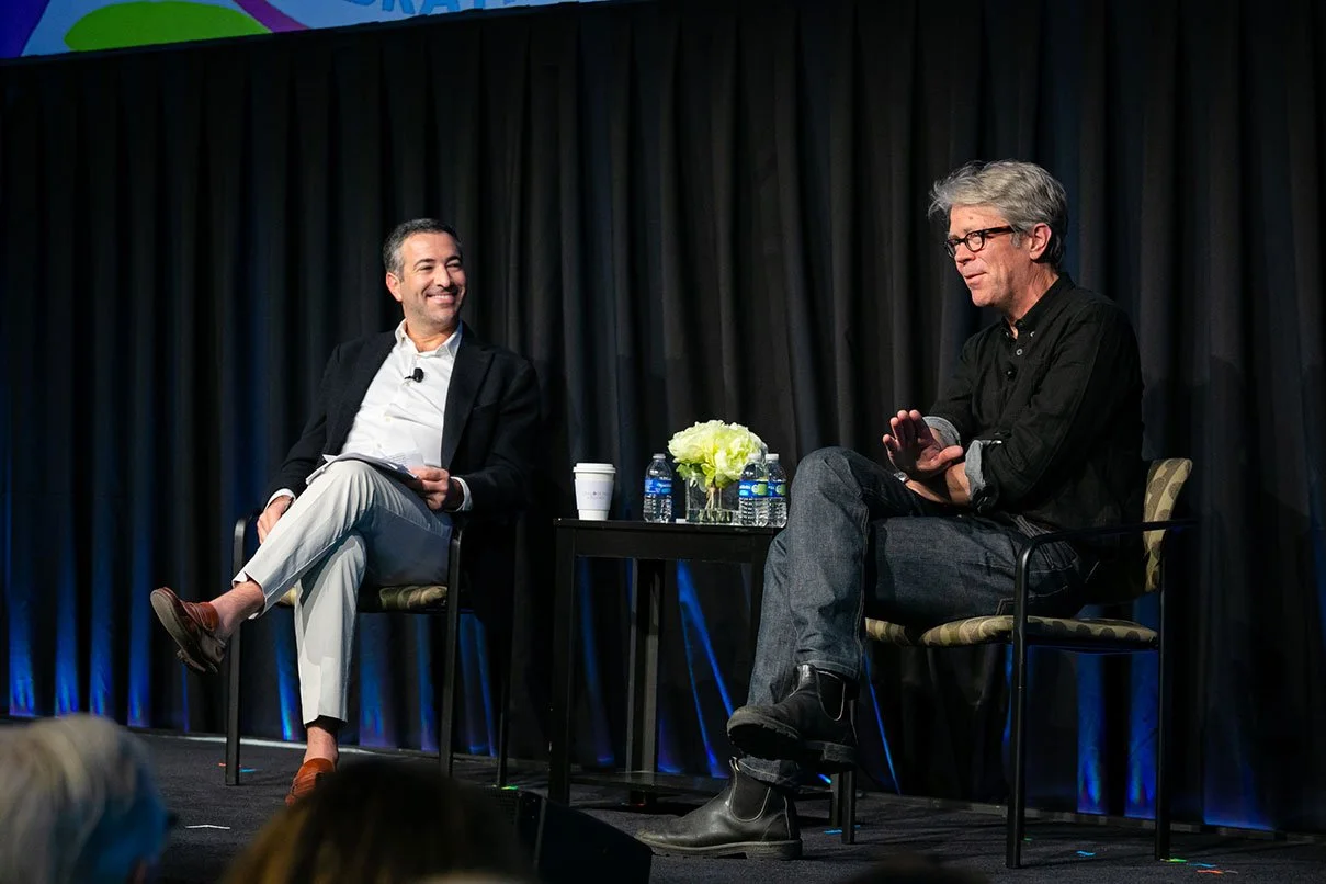 Two men sitting on stage in chairs, engaged in conversation, with a small table between them holding water bottles, a coffee cup, and a floral arrangement, against a dark curtain background.