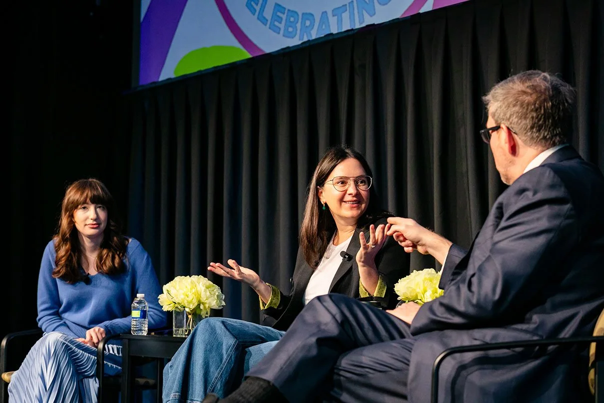 Three people seated on stage in a panel discussion, with black curtains and a colorful banner in the background.