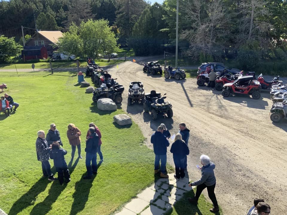 A group of people standing on a grassy area near parked ATVs and off-road vehicles on a dirt path with trees in the background.