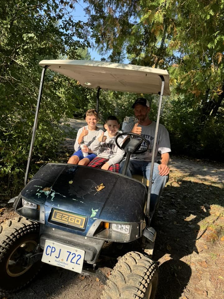 Three people sitting in a golf cart on a wooded path, giving thumbs up.