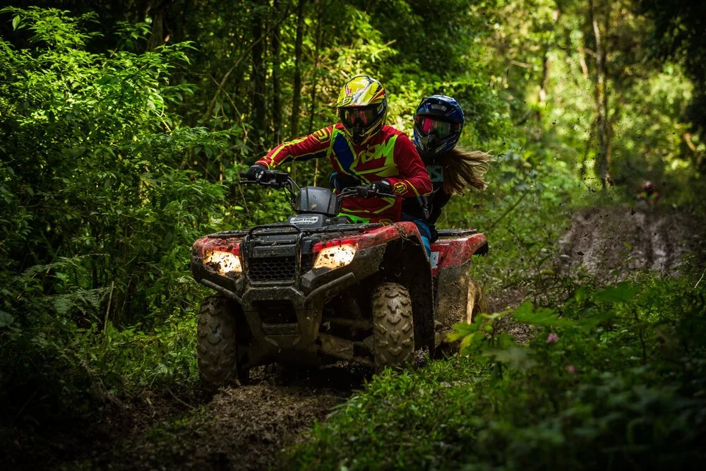 Two people riding a red ATV through a forest trail, wearing helmets and protective gear.