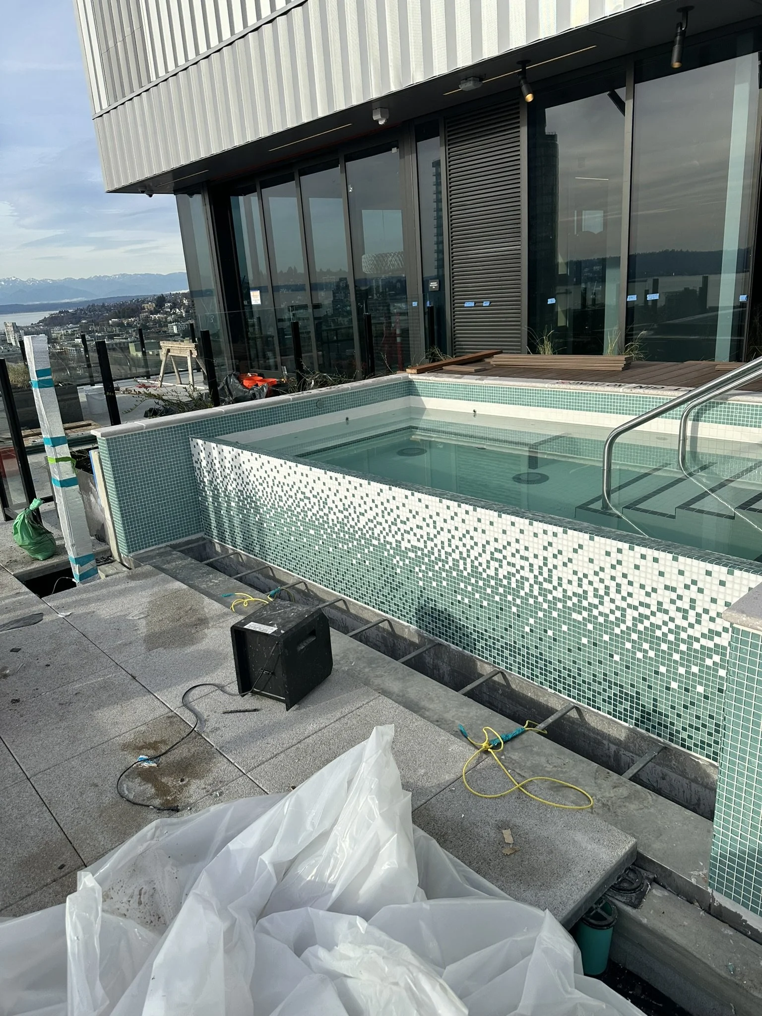 A rooftop hot tub under construction with green mosaic tiles, in front of a modern building with floor-to-ceiling windows, overlooking a cityscape with mountains in the distance.