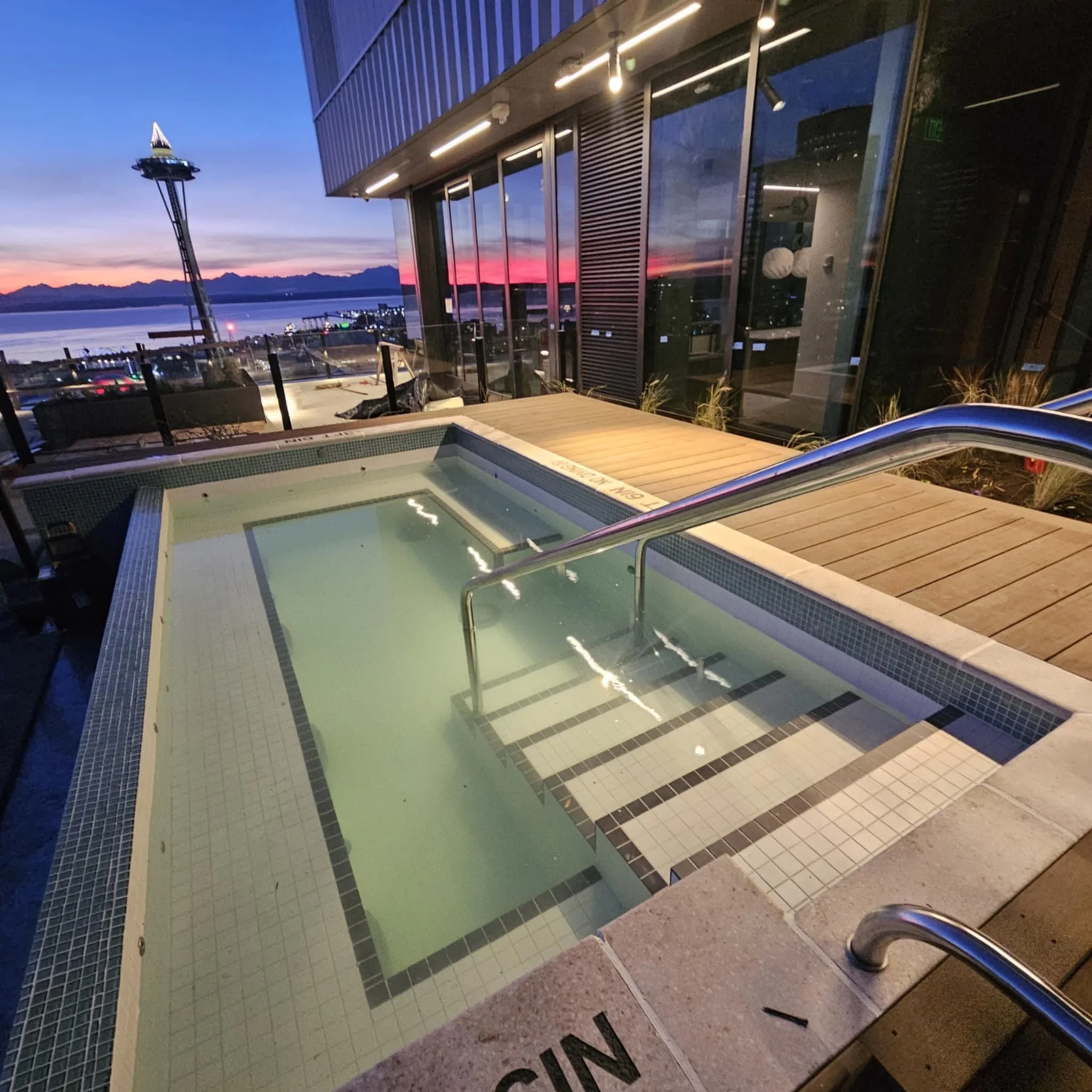 Outdoor hot tub with steps and metal handrail, next to a wooden deck and glass walls, overlooking a sunset over water with the Seattle Space Needle in the distance.