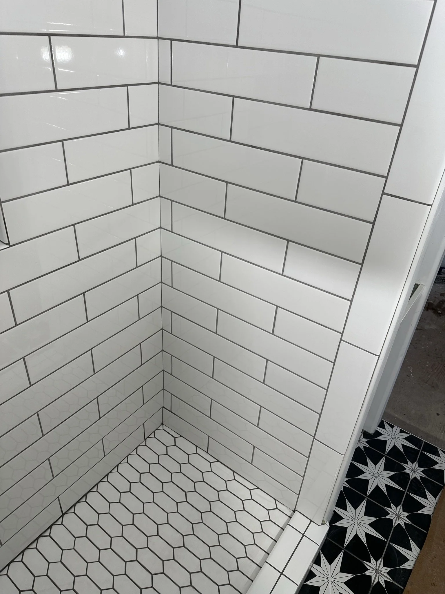 Interior corner of a tiled bathroom shower with white subway tiles and black grout on walls, white hexagonal tiles on the floor with black grout, and a black and white patterned tile on the floor outside the shower.