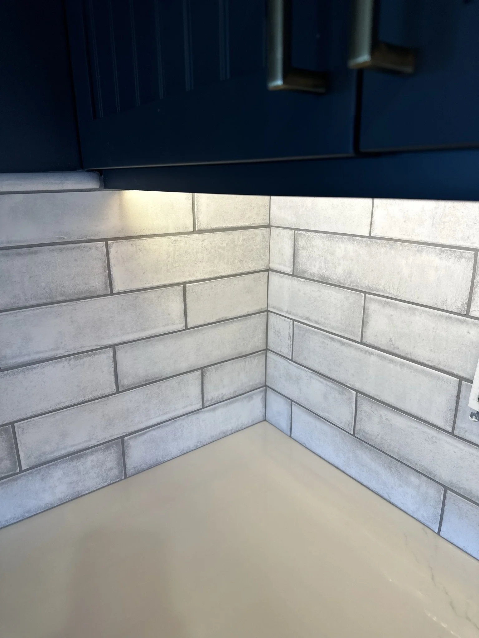 Close-up of a corner in a kitchen with light gray brick tiles on the wall, a beige countertop, and dark upper cabinets with metallic handles.