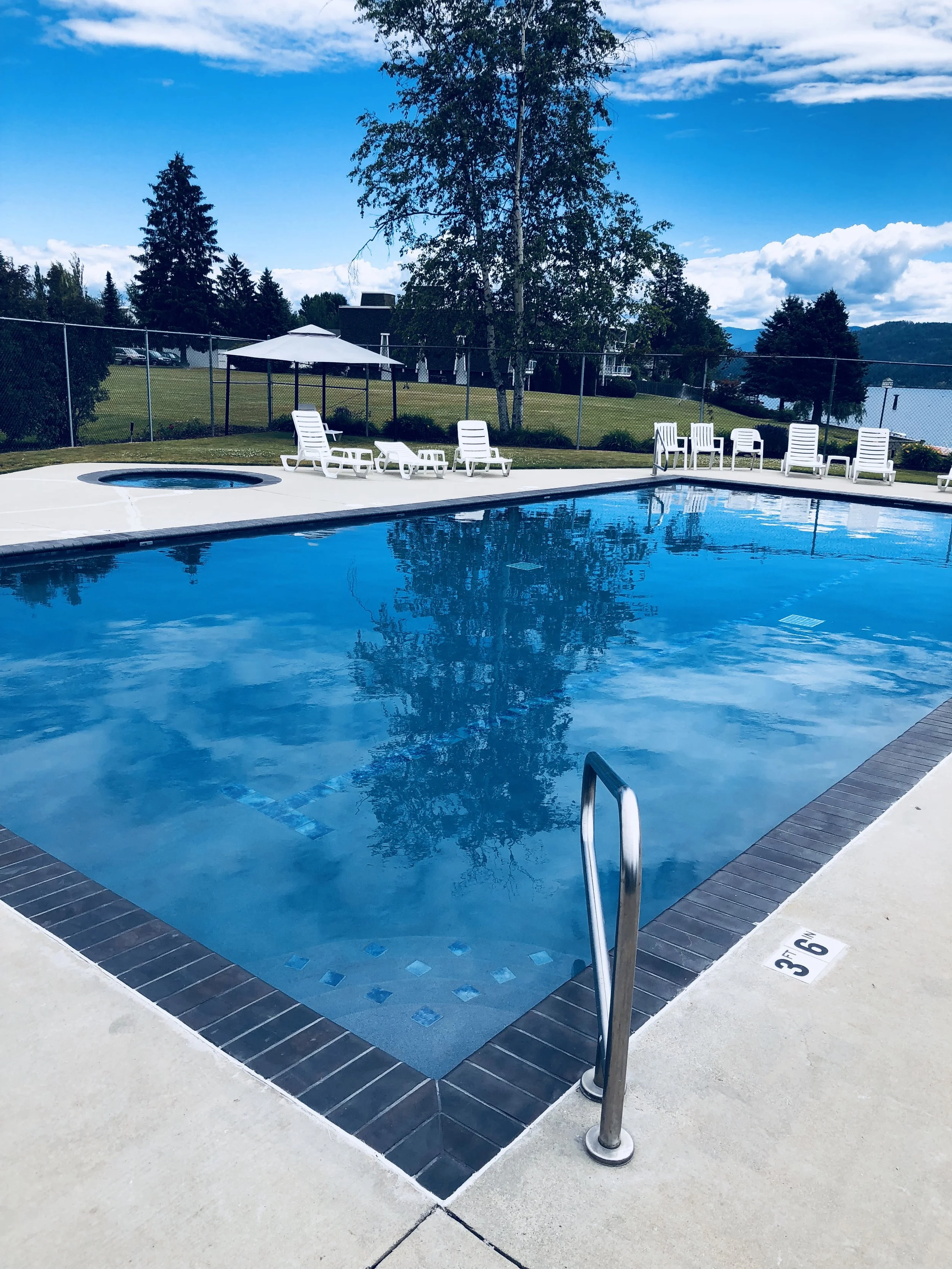 Outdoor swimming pool with ladders, surrounded by white lounge chairs, on a sunny day with trees and buildings in the background.