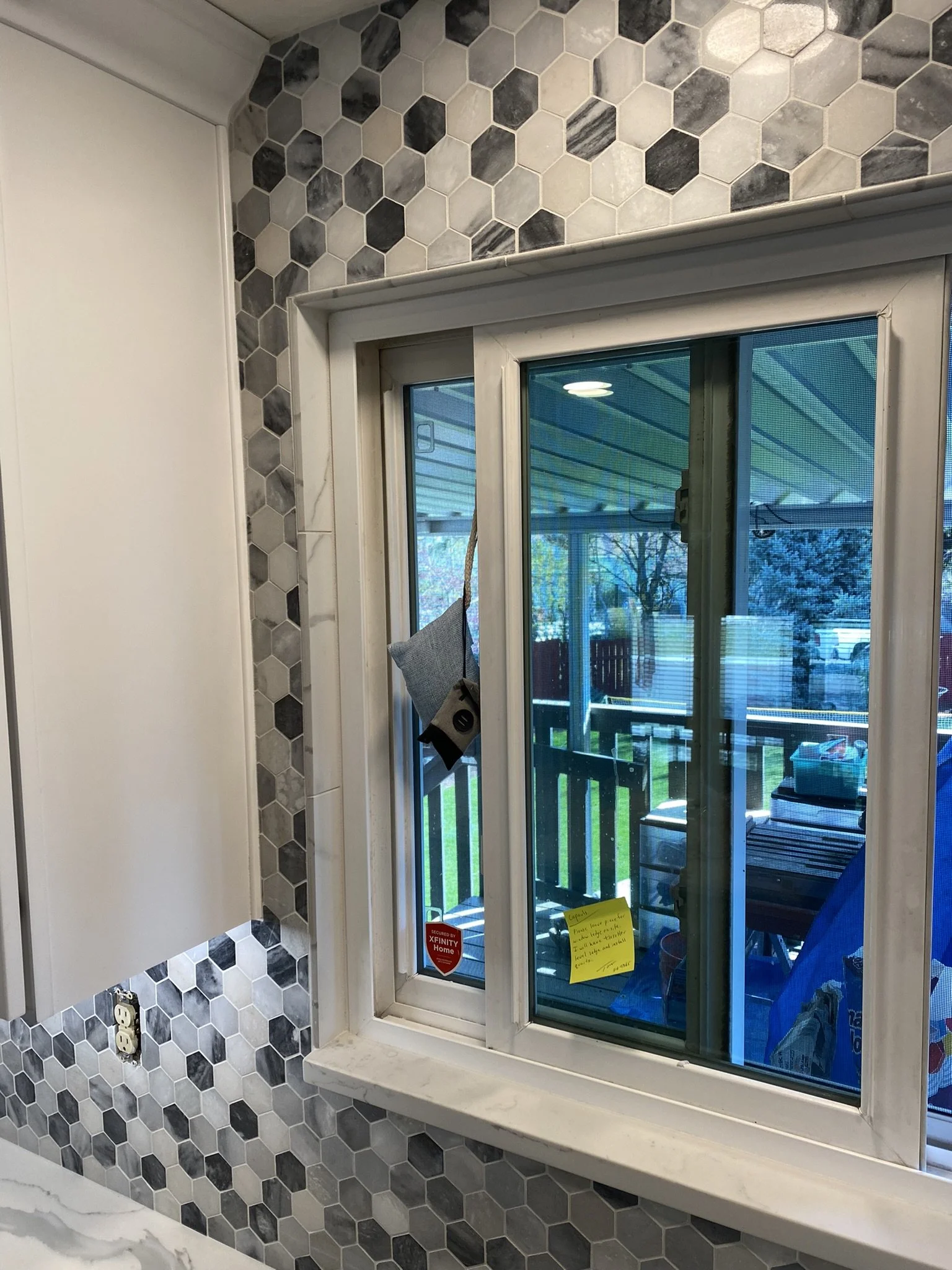 A kitchen window with a white frame, overlooking a porch with outdoor furniture. The wall around the window is decorated with gray and white hexagon tiles, and there is a white cabinet and outlet nearby.