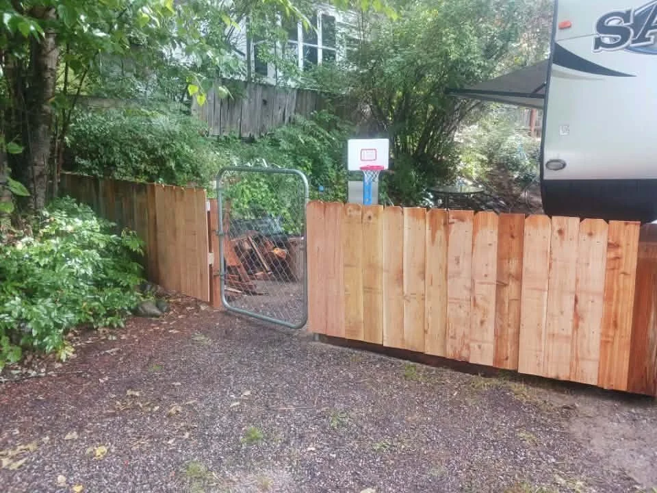 A backyard with a wooden fence, a small gate, and a basketball hoop in the background.