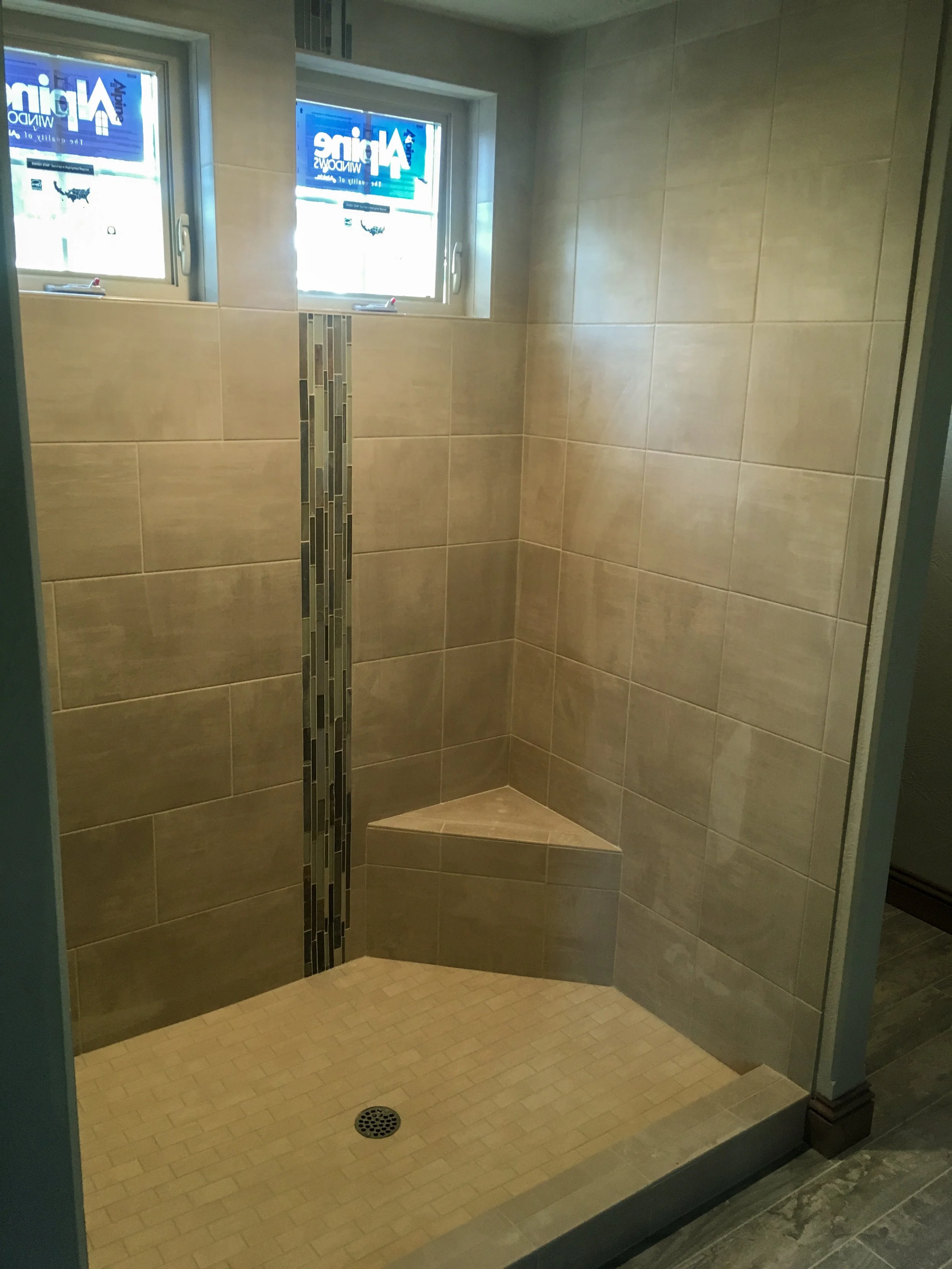 A walk-in shower with beige tiles, a vertical strip of decorative tiles, a small corner seat, and two small windows at the top with blue signs.