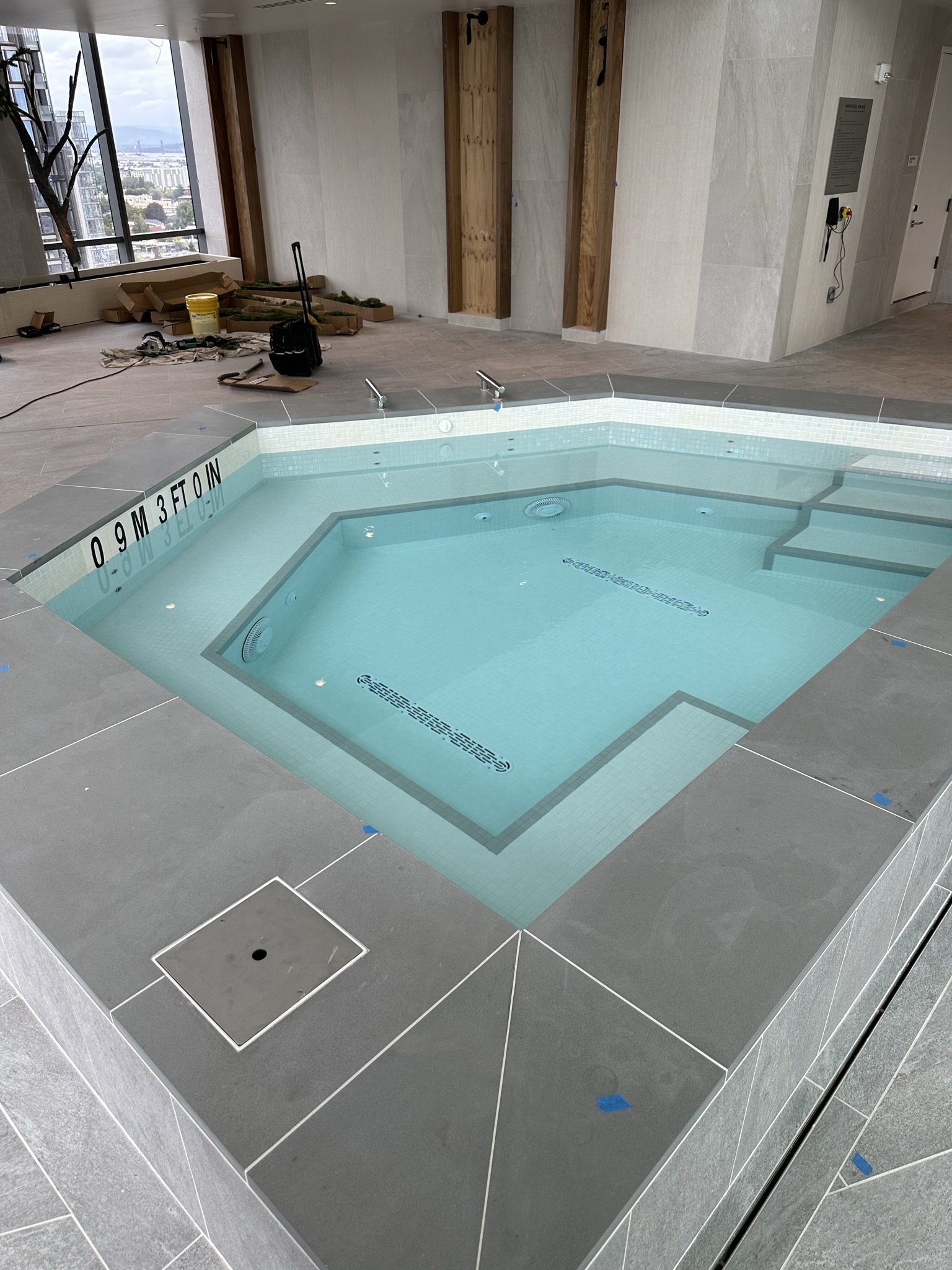 Indoor pool with a glass wall view of a city, surrounded by gray tiles and beige walls, with construction materials and tools nearby.