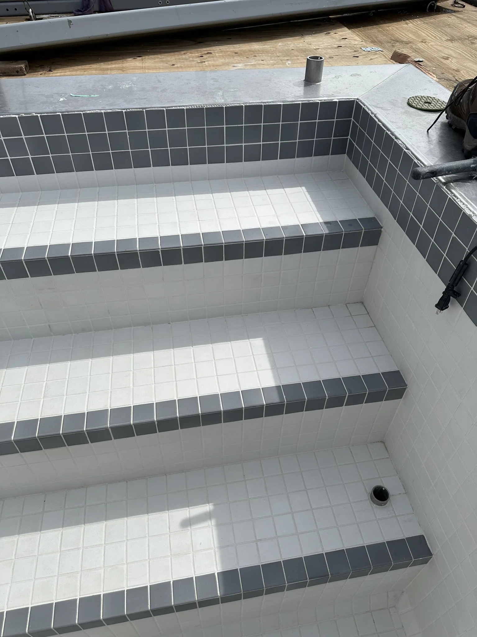 In progress of building a tile-lined hot tub with three steps, featuring white and gray tiles, on a construction site with wooden decking and some tools around.