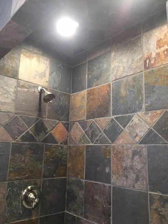 Close-up of a slate tile shower with a metal showerhead and a round water control knob, with a ceiling light illuminating the shower area.
