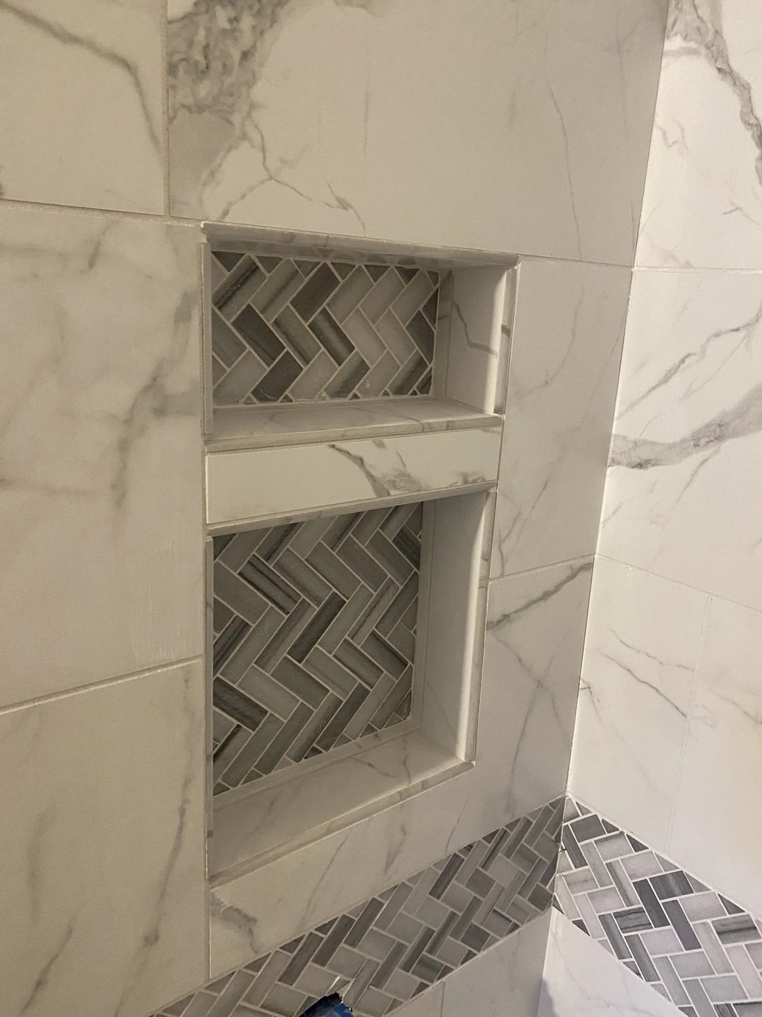 Close-up of a shower wall with marble-like tiles and a built-in shampoo niche featuring herringbone-patterned tiles.