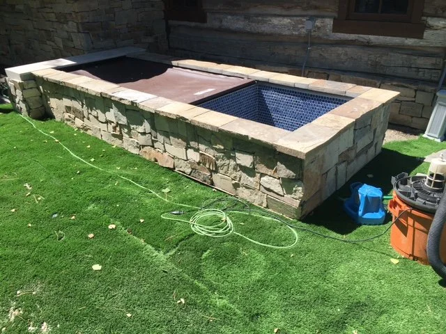 Stone hot tub with wooden cover on a grassy lawn, with garden tools and a nearby window.