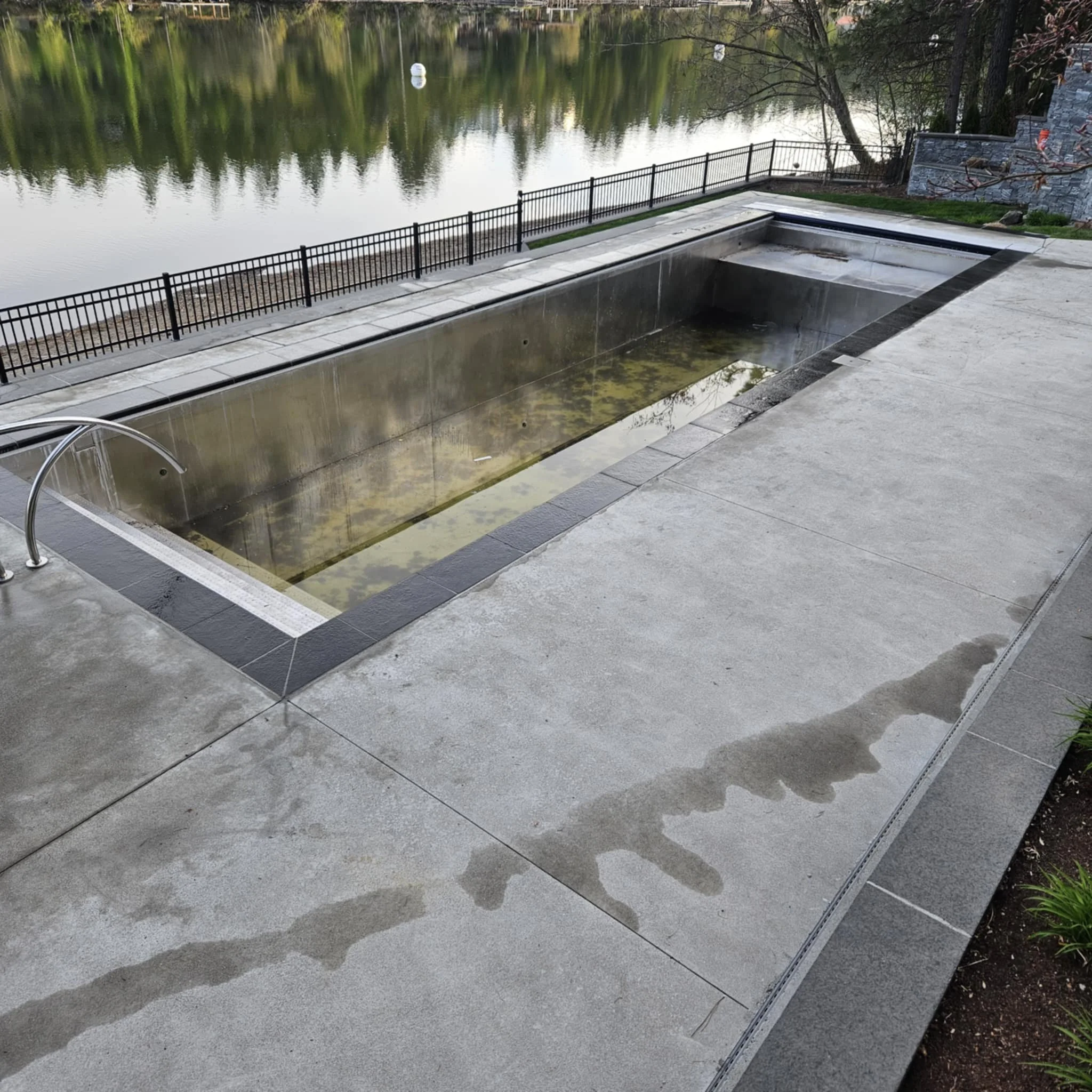 Empty rectangular outdoor pool with a waterline that has algae and debris, surrounded by a concrete deck, with a lake and trees in the background.