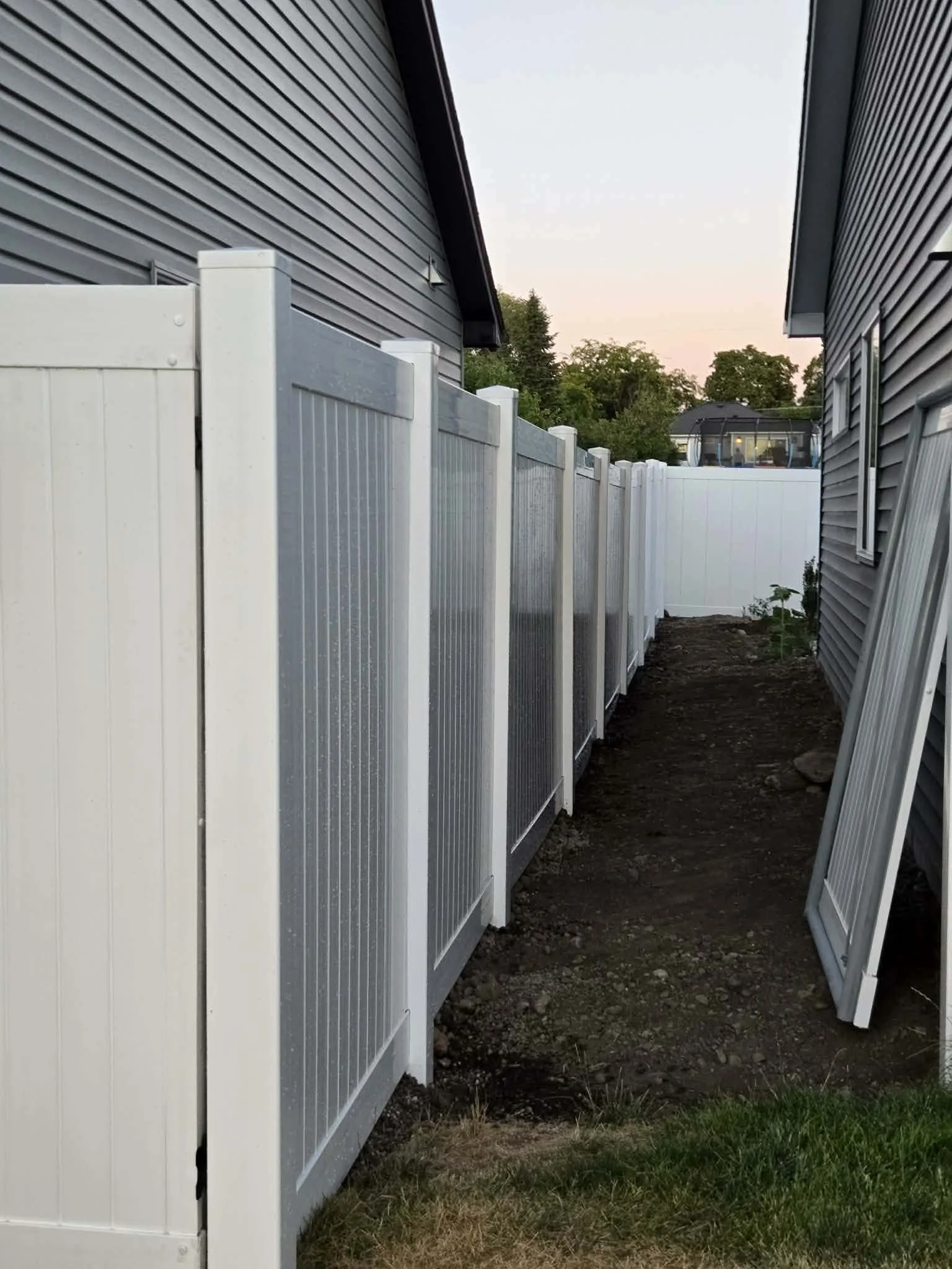 Residential backyard with white privacy fence, gray house siding, and a dirt pathway between the fence and house, with some plants at the far end, during sunset or early evening.