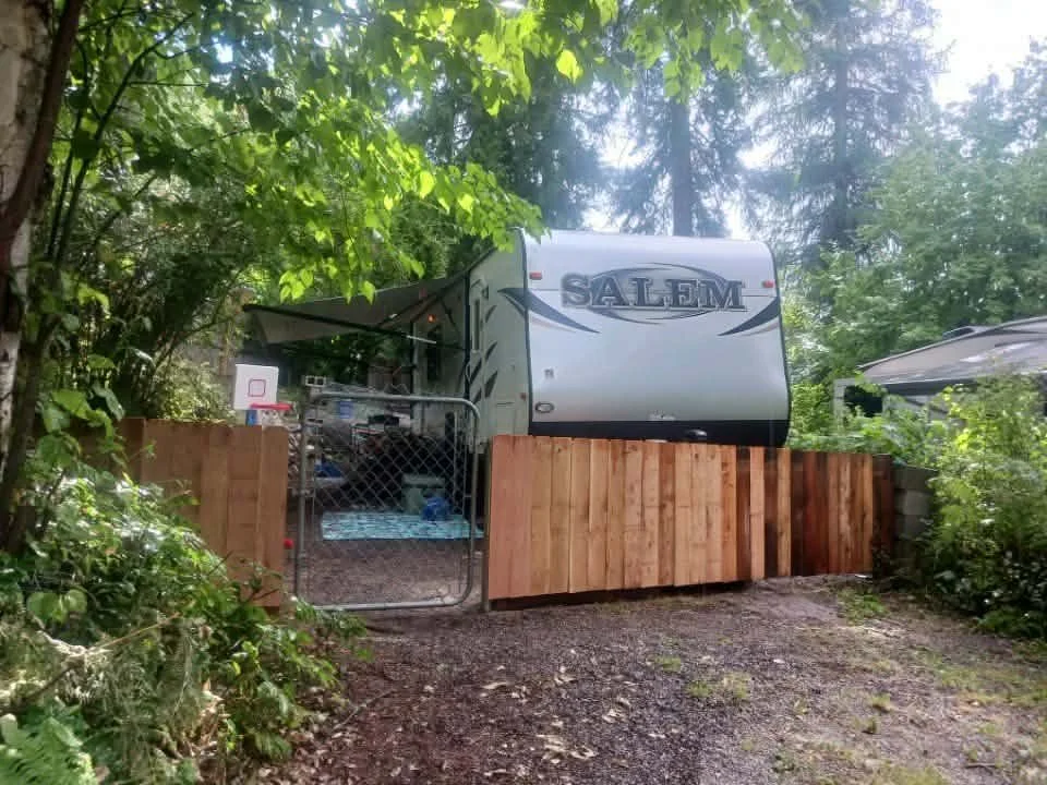A travel trailer labeled 'Salem' parked behind a wooden fence, with a gate, in a wooded area.