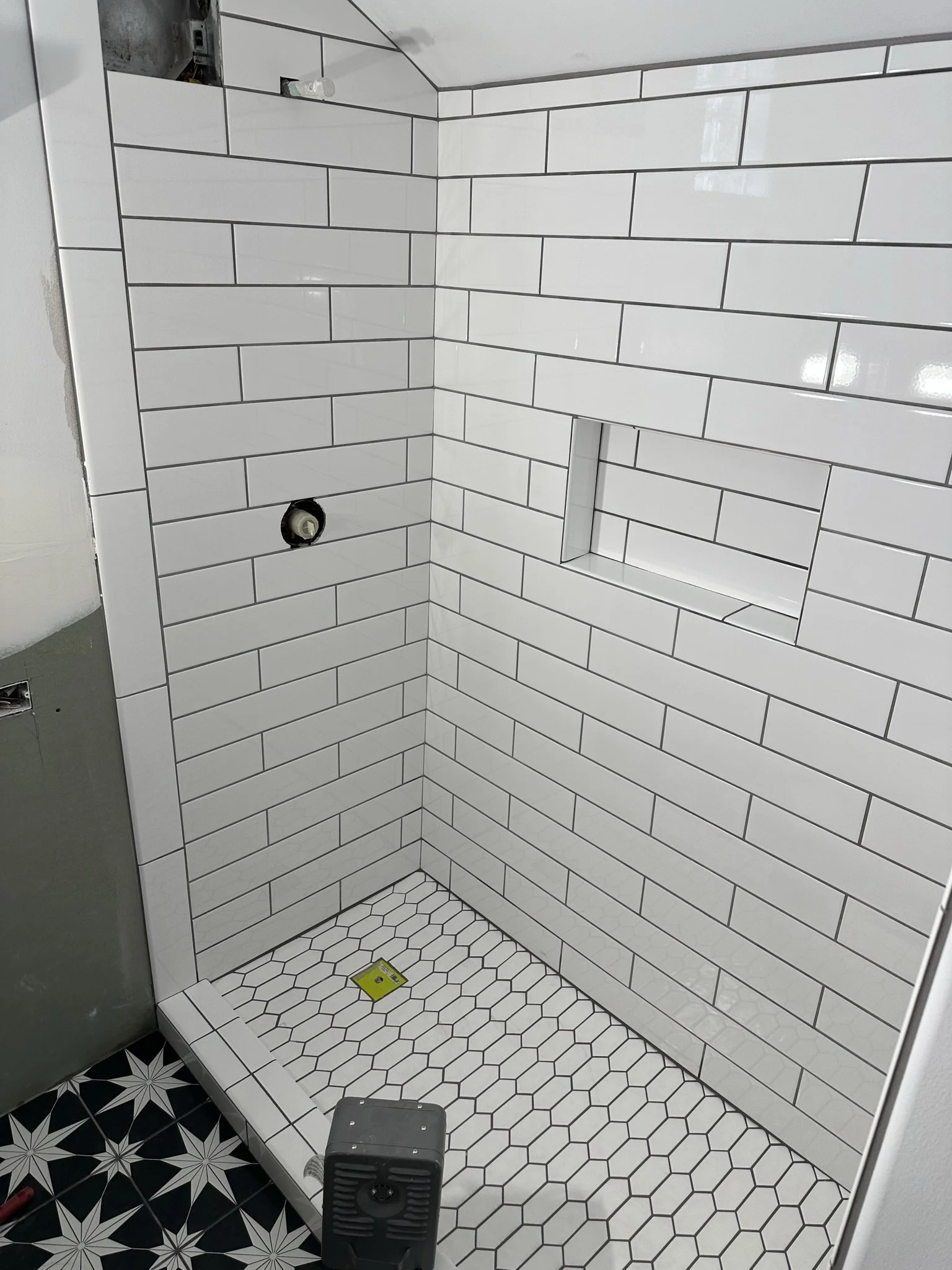 A partially constructed shower with white subway tile walls, a built-in niche, hexagon tile flooring, and black and white patterned floor tiles outside of the shower.