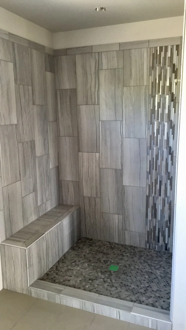 A walk-in shower with beige and gray marbled tiles on the walls and a pebble stone floor. There is a built-in bench in the corner and a vertical strip of small, rectangular mosaic tiles on the right wall.