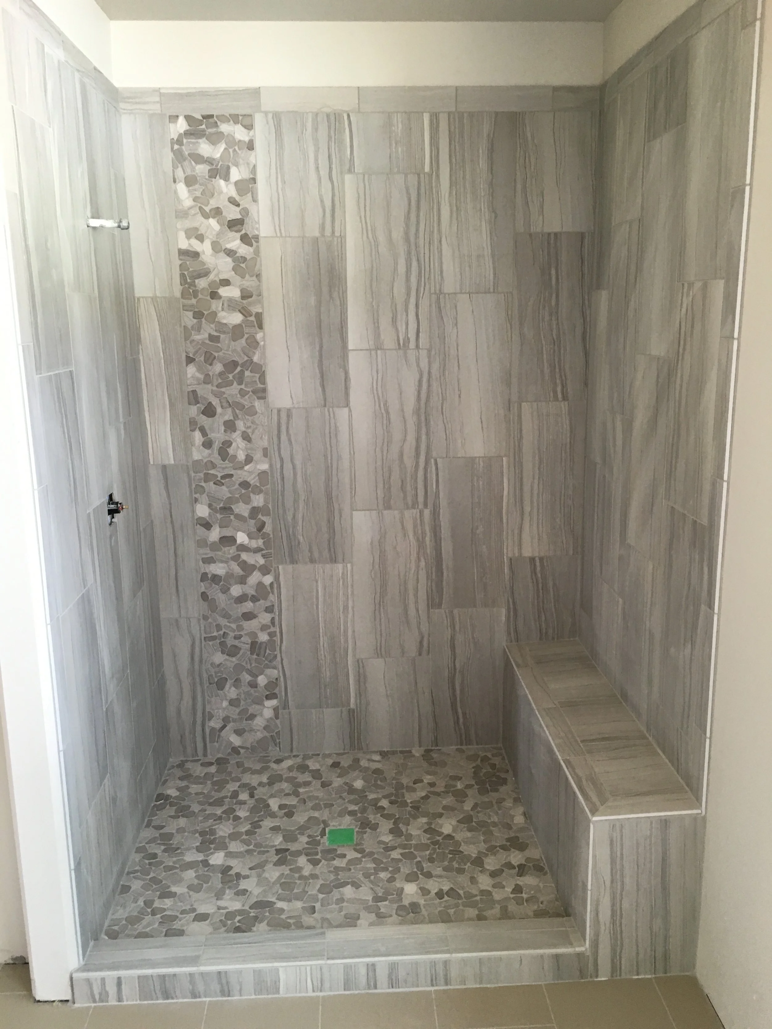 Empty shower with gray tiles and a pebble stone accent strip on the wall and floor, including a built-in bench.