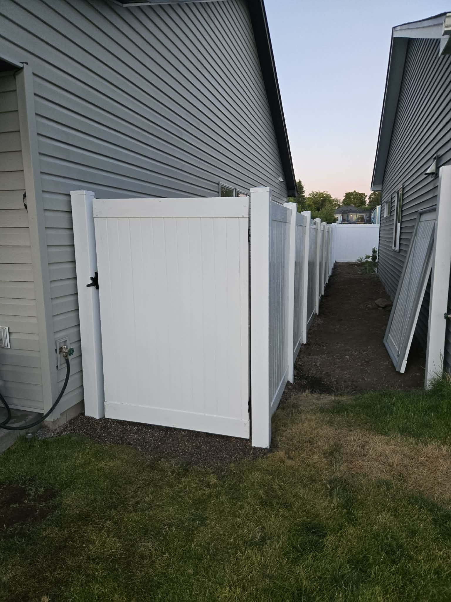 White vinyl fence panels forming a pathway between two houses with gray siding, one panel leaning against the house on the right, and the grass and dirt ground.