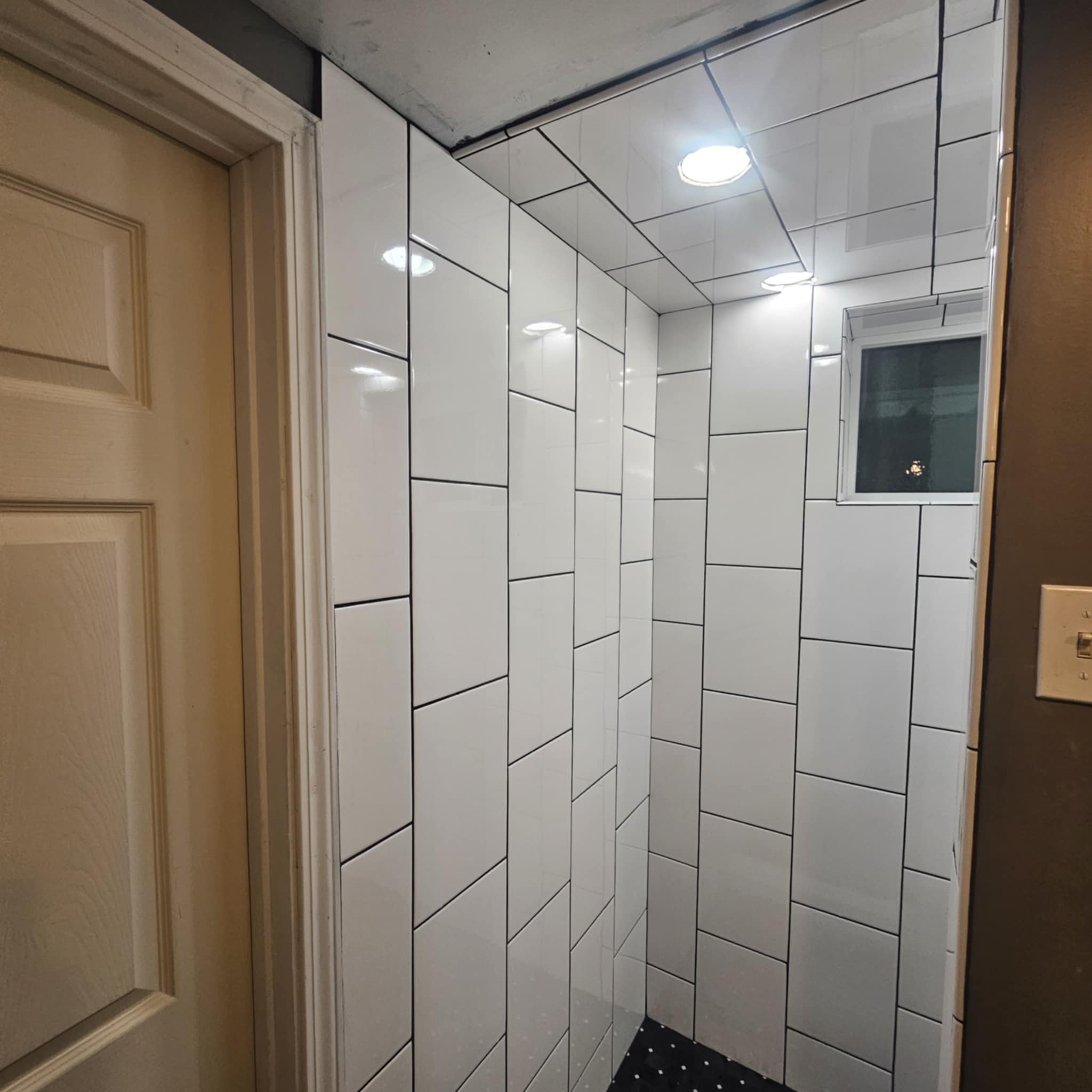 A corner of a bathroom with large white subway tiles on the walls and a small window. The ceiling has recessed lighting, and there is a beige door on the left side.