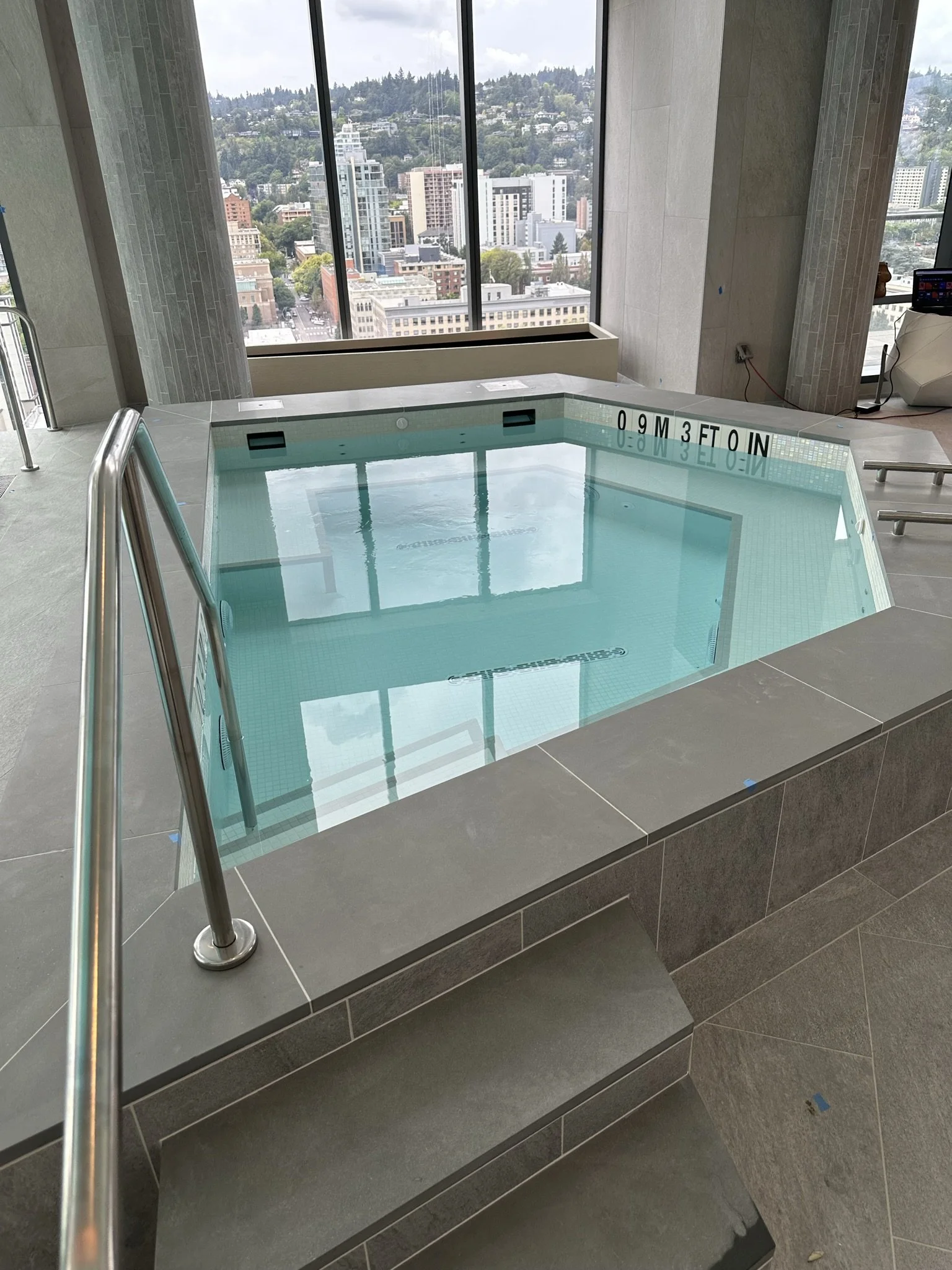 Indoor rooftop spa with steps leading into the water, large windows showing cityscape and hills, stainless steel handrail, and safety depth markings on the side.