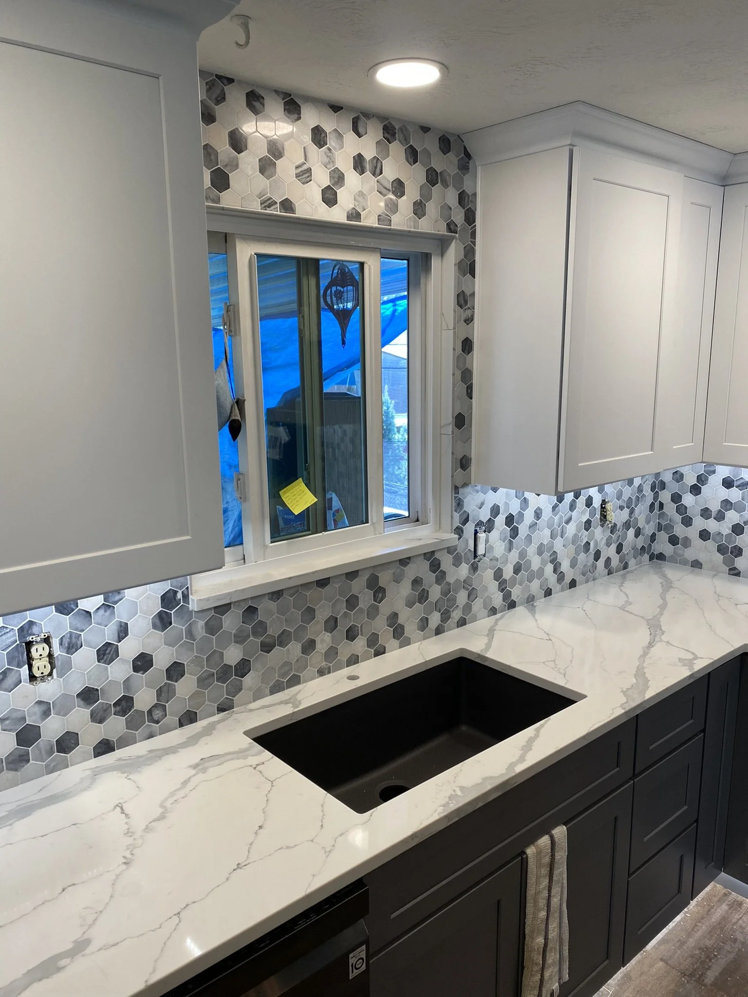 Modern kitchen with white upper cabinets, black lower cabinets, a white marble countertop, hexagon gray backsplash tiles, a window with blue covering, and a black sink.