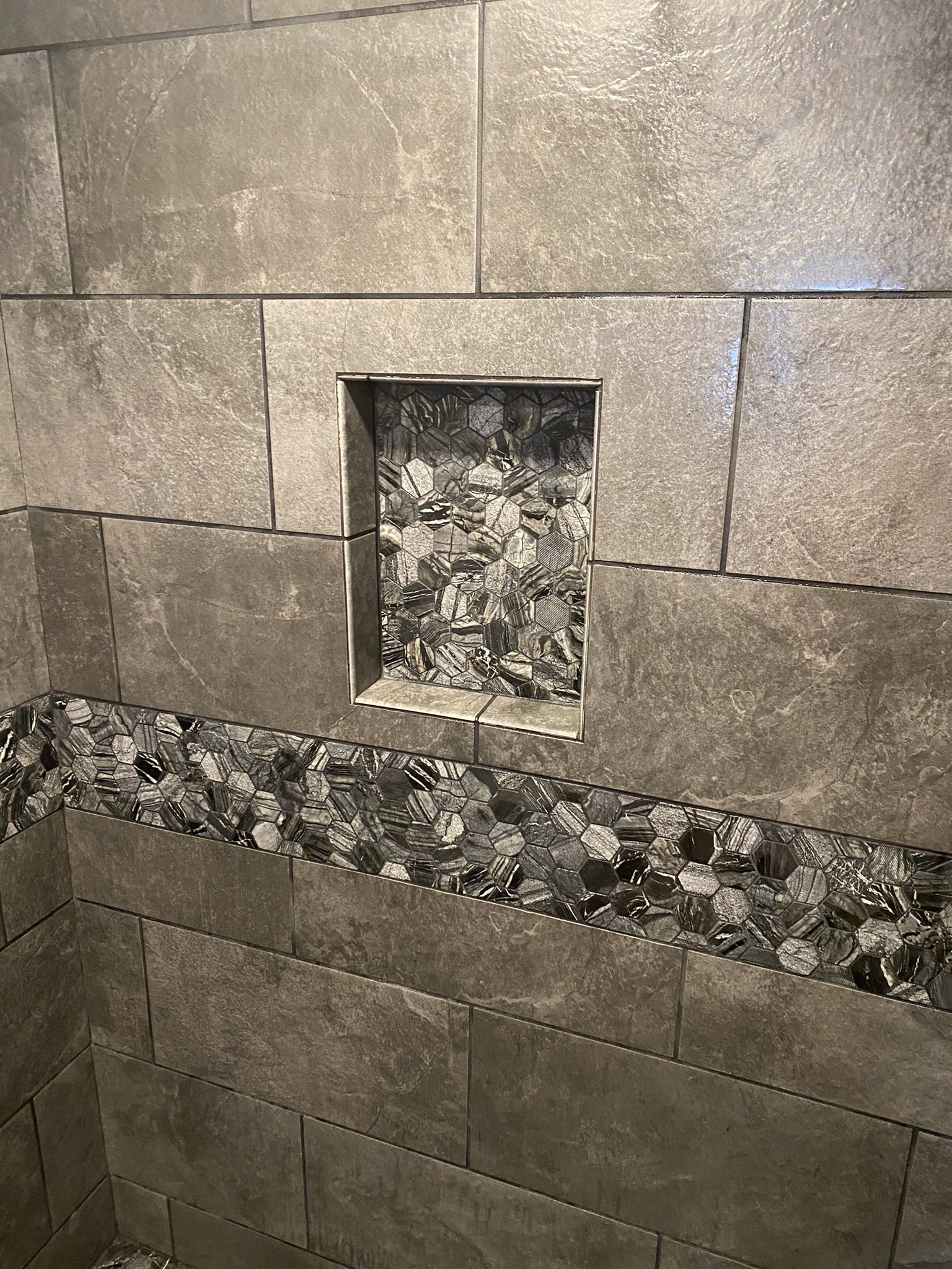 Close-up of a tiled wall in a shower with a built-in shelf made of hexagonal mosaic tiles, surrounded by large gray rectangular tiles.